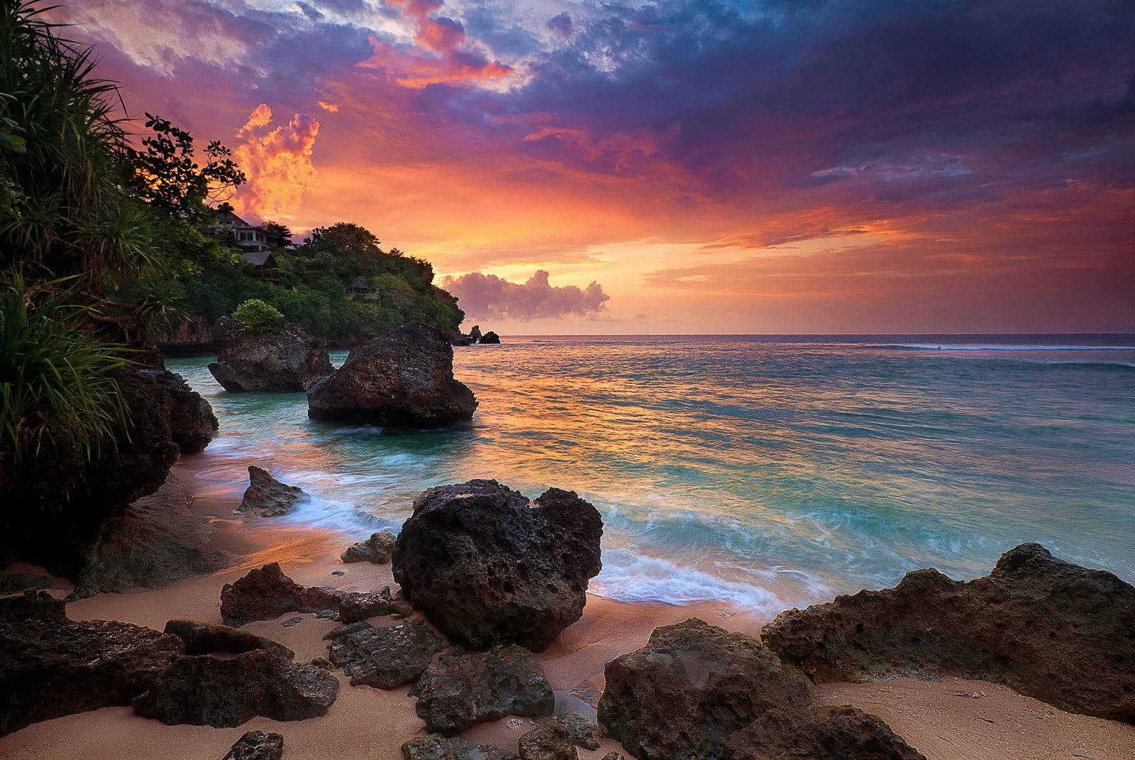海灘 桌布 壁紙 — 一輪充滿活力的夕陽，在岩石熱帶海灘和碧綠海浪之上，將天空染成火熱的橙色和紫色。