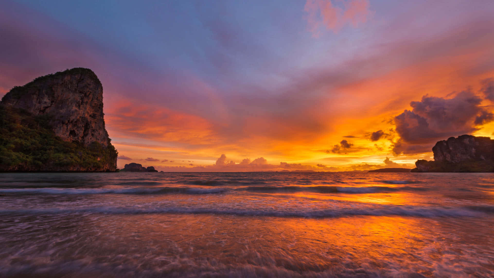 Beach Sunset HD 桌布 壁紙 — 一輪充滿活力的夕陽將天空染成火熱的橙色和紫色色調，映照在兩側有岩石懸崖的平靜海洋上。