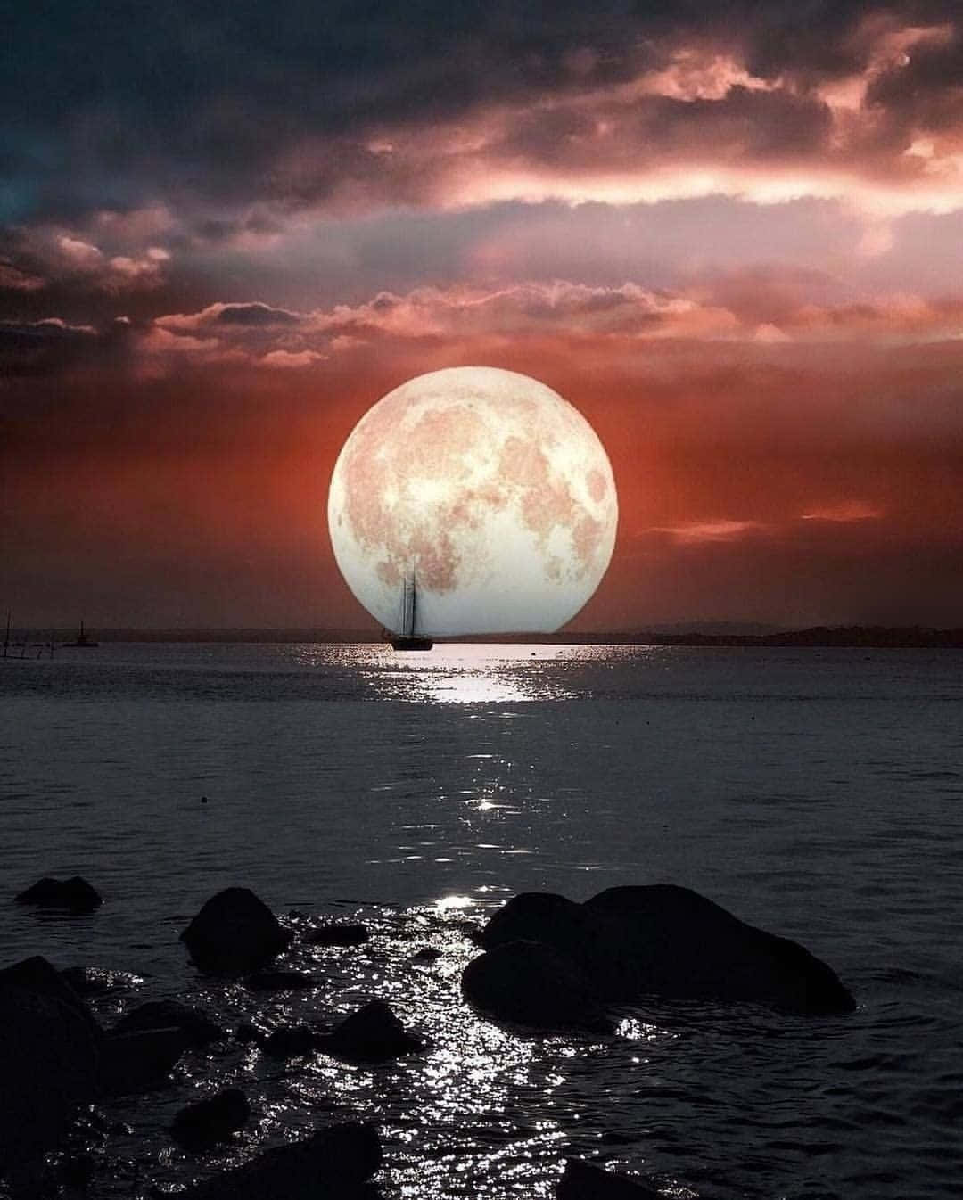 Beautiful Moon 桌布 壁紙 — 一輪巨大的發光月亮在平靜的海面上升起，下方有一艘帆船的剪影，前景是深色的岩石。