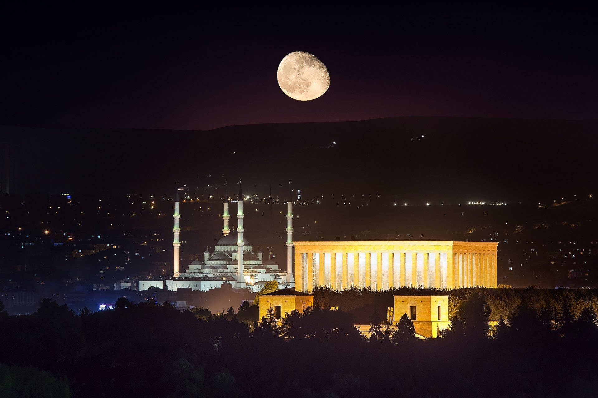 Ankara 桌布 壁紙 — 一輪明亮的彎月懸掛在一座宏偉的燈火通明的清真寺和現代建築之上，它們坐落於夜晚黑暗的森林山谷中。
