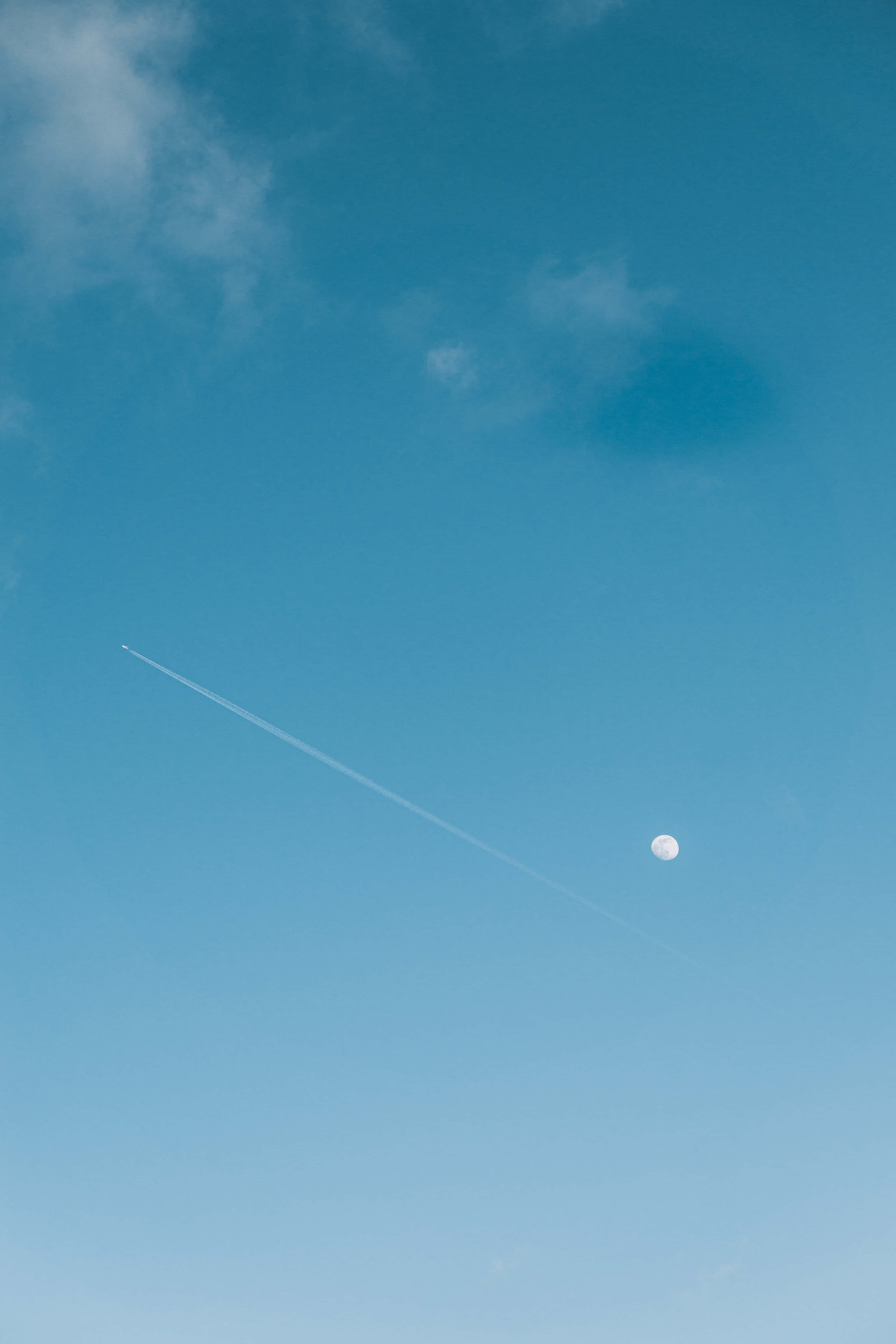 月光 桌布 壁紙 — 一輪明亮的月亮在長長的飛機雲旁邊發光，飛機雲劃過廣闊的藍天，天空中有散落的雲朵。