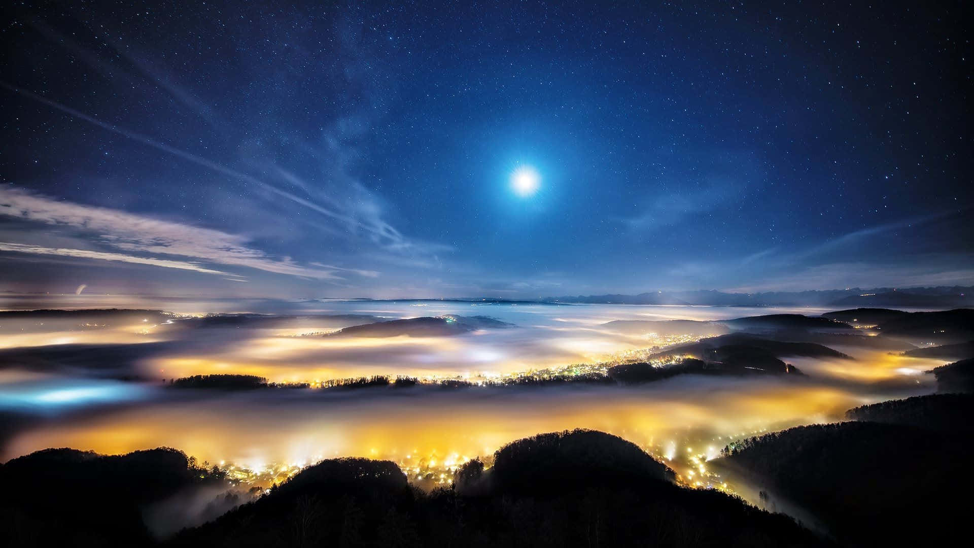 夜空 桌布 壁紙 — 一輪明亮的月亮照耀在黑暗森林山丘之間的發光雲海之上，下方是星空夜空。