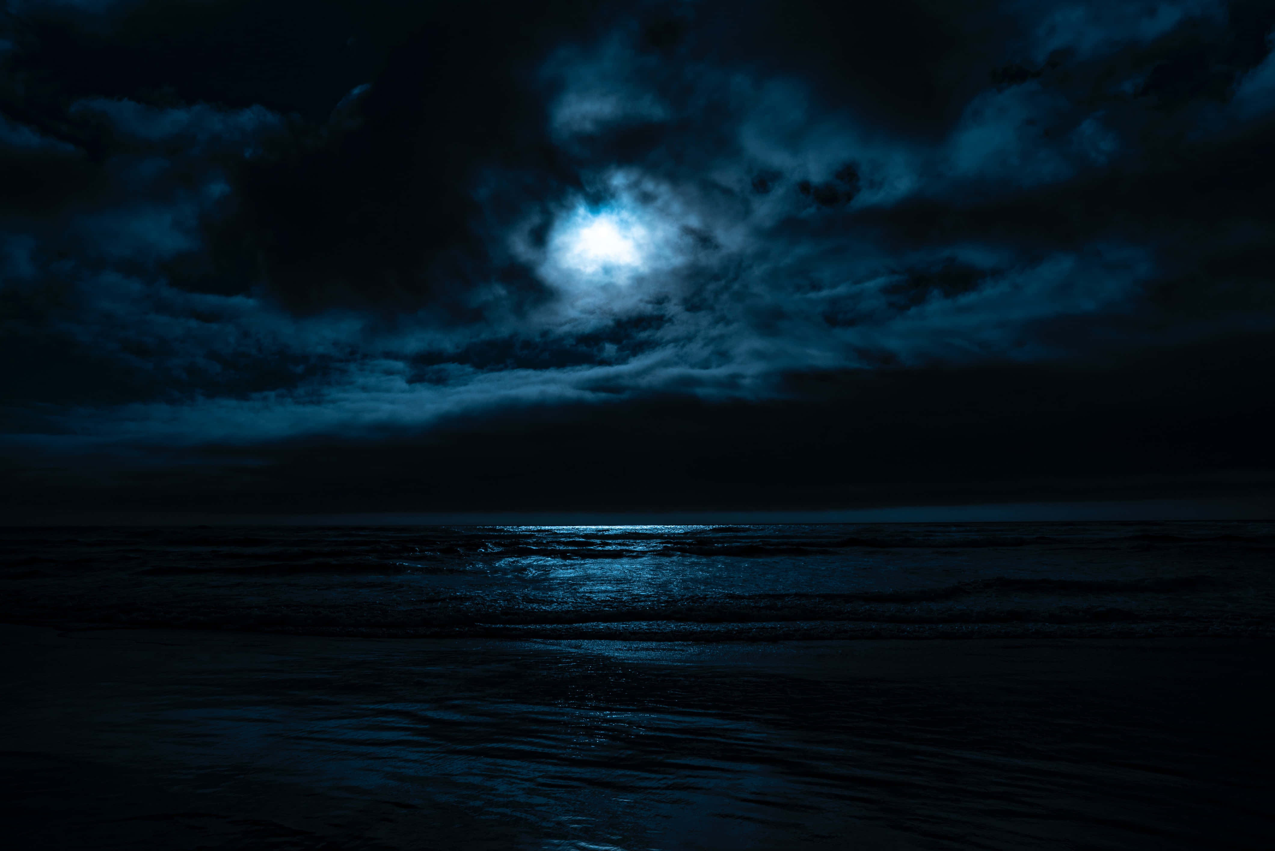 Dark Ocean 桌布 壁紙 — 一輪明亮的月亮穿透黑暗的暴風雲，在夜晚不安的海洋上投下空靈的藍色光芒。