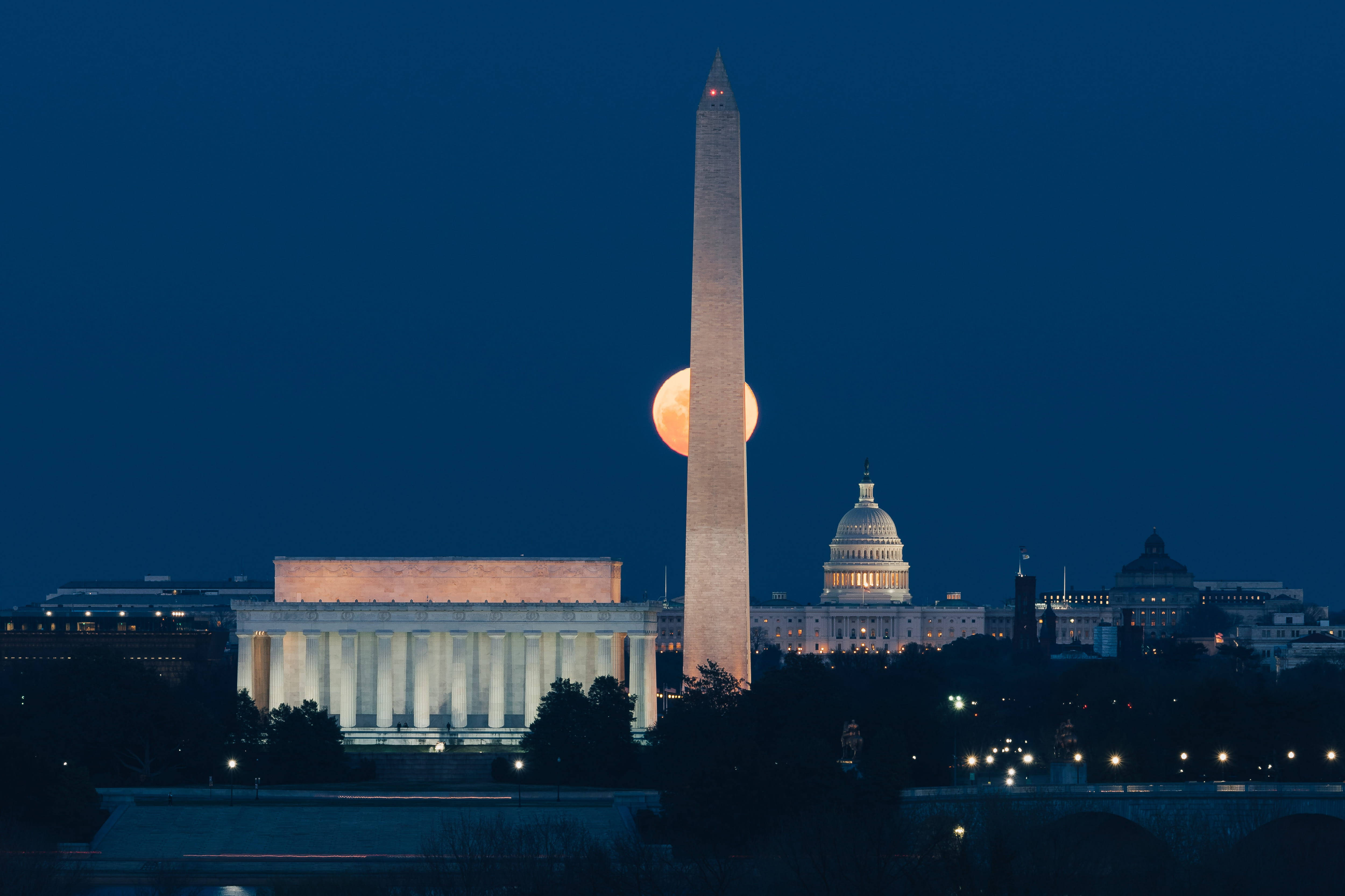 Washington 桌布 壁紙 — 一輪明亮的橙色月亮在華盛頓紀念碑後升起，林肯紀念堂和國會大廈在暮色天空下閃耀。