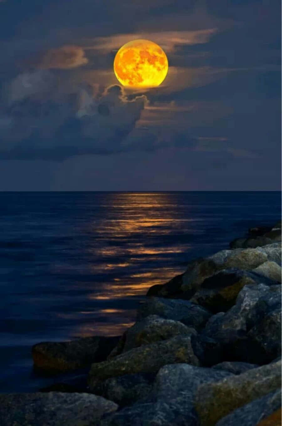 Beautiful Moon 桌布 壁紙 — 一輪明亮的橙色月亮在黑暗的海洋上方發光，在崎嶇的岩石旁的水面上投下金色的倒影。