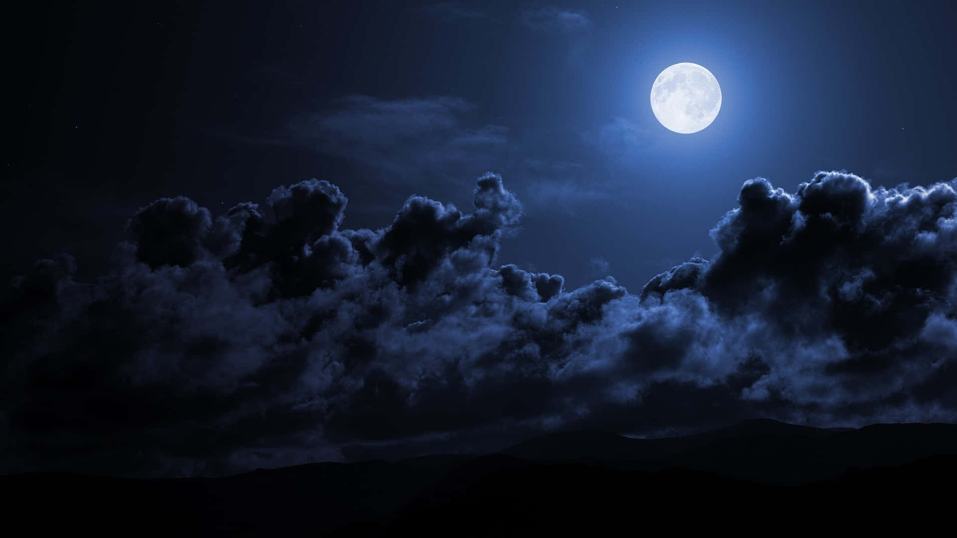 月光 桌布 壁紙 — 一輪明亮的滿月在夜間將冷藍色的光芒投射在戲劇性的暴風雲上，這些雲在黑暗的山脈背景下形成剪影。