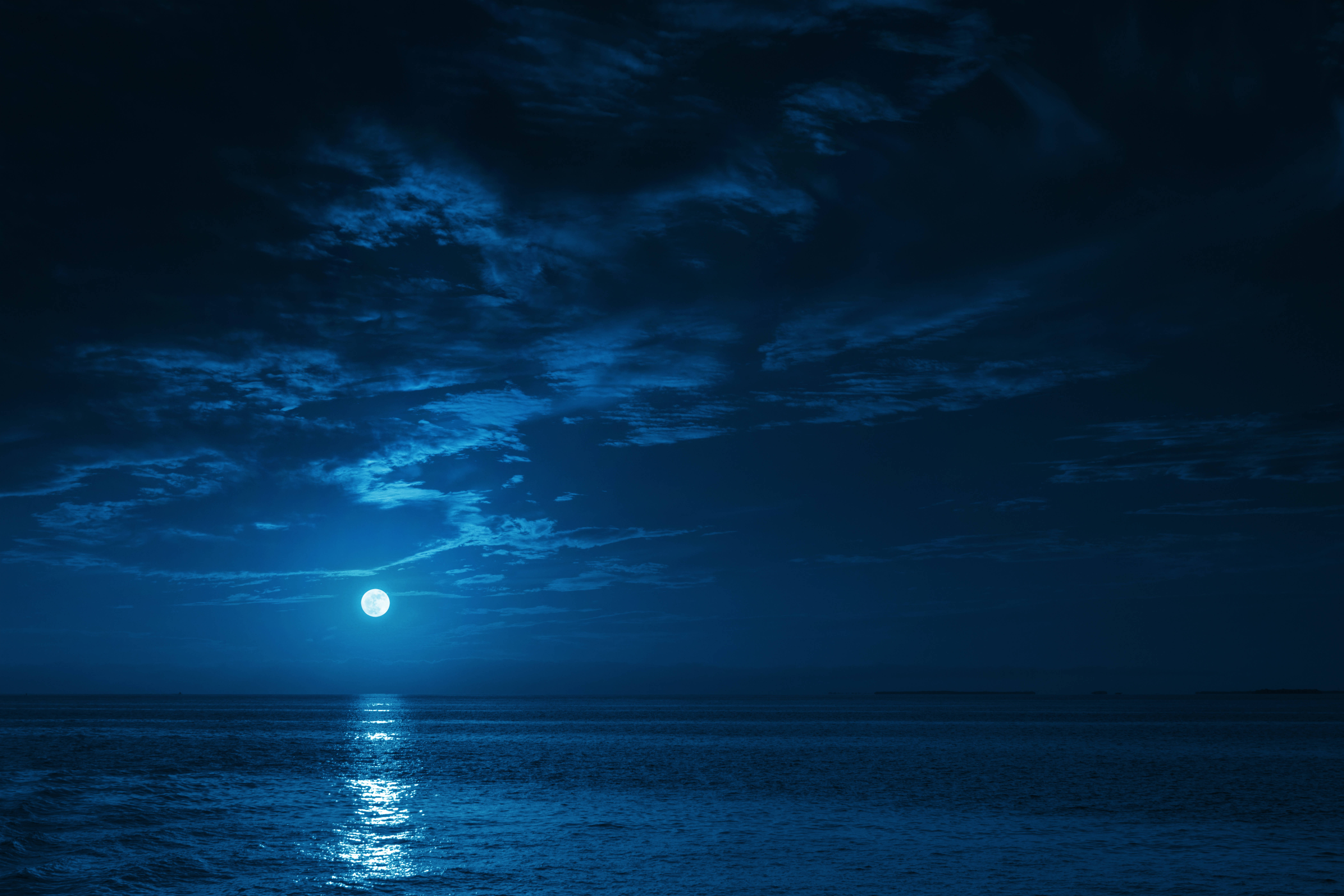 夜空 桌布 壁紙 — 一輪明亮的滿月在戲劇性的多雲夜空下，在平靜的海洋上投射出寧靜的藍色光芒。