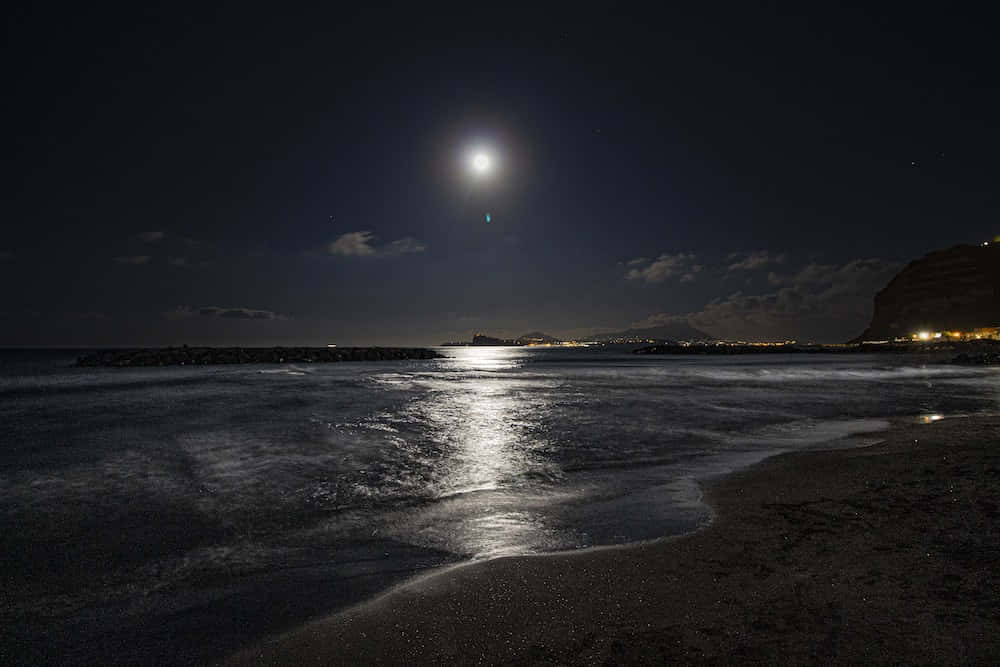 海灘 桌布 壁紙 — 一輪明亮的滿月在星空下，在寧靜的夜晚海灘上投下一條閃爍的光路，遠處有城市燈光和黑暗的懸崖。