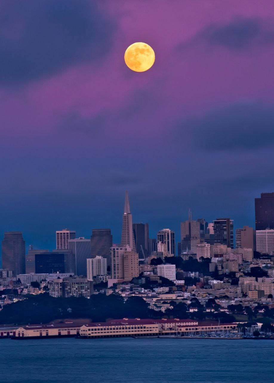 san francisco iPhone 桌布 壁紙 — 一輪明亮的滿月在暮色中照耀著舊金山的天際線，在雲層和城市景觀上投下紫色調。
