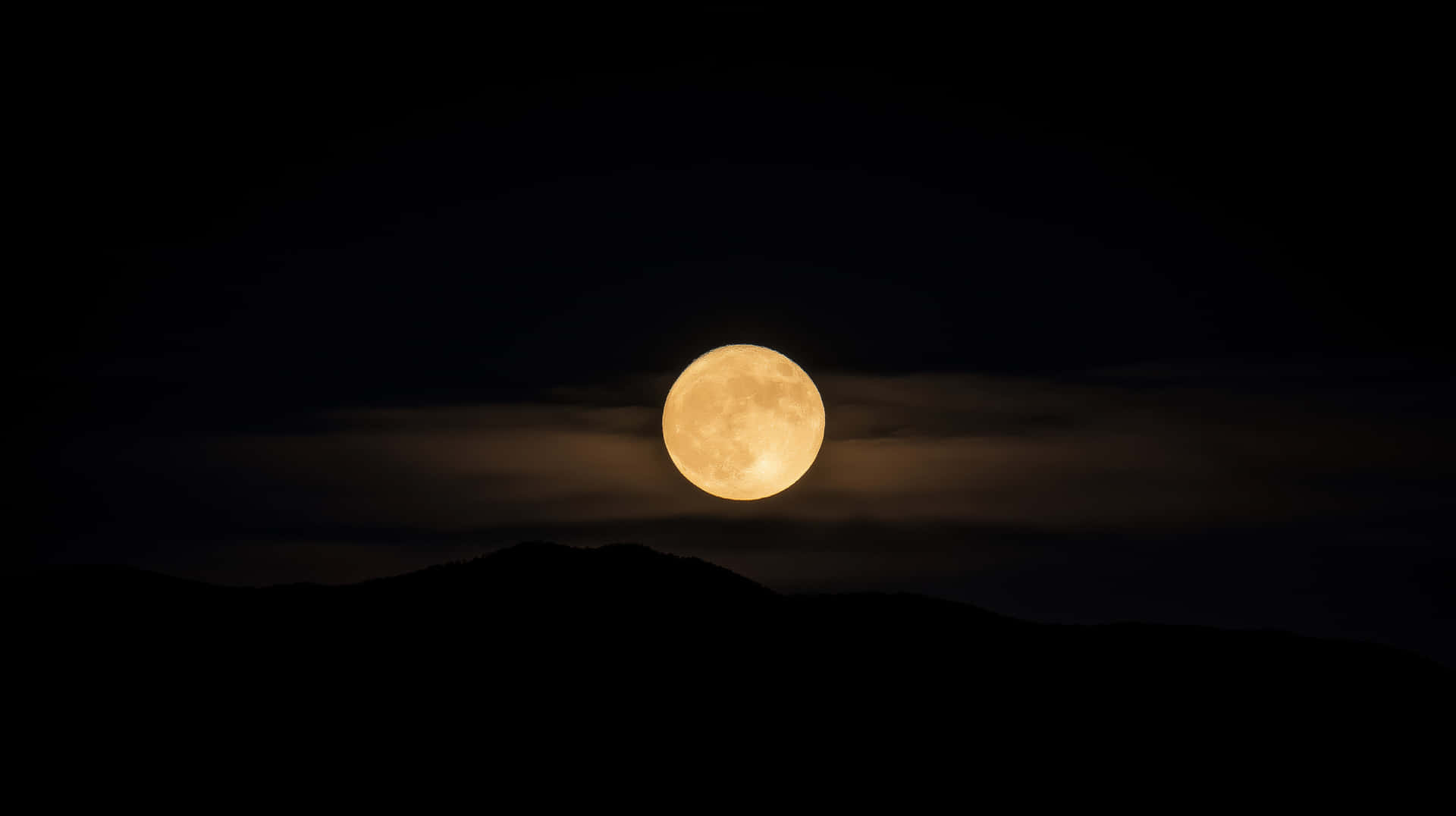Golden Full Moon Rising Above Silhouetted Mountains in the Night Sky 壁紙 — 一輪明亮的滿月散發琥珀色光芒，懸掛在陰雲密布的夜空下黑暗山巒的剪影之上。