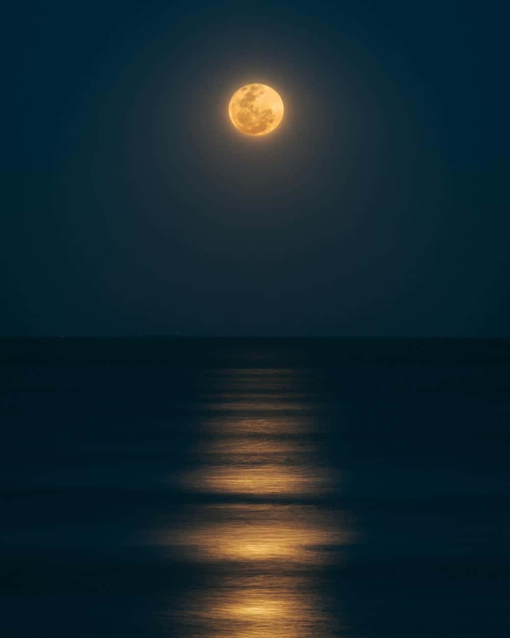 Moonlight 桌布 壁紙 — 一輪明亮的滿月散發琥珀色光芒，照耀著平靜的海洋，在黑暗的水面上投射出一條閃爍的光路。