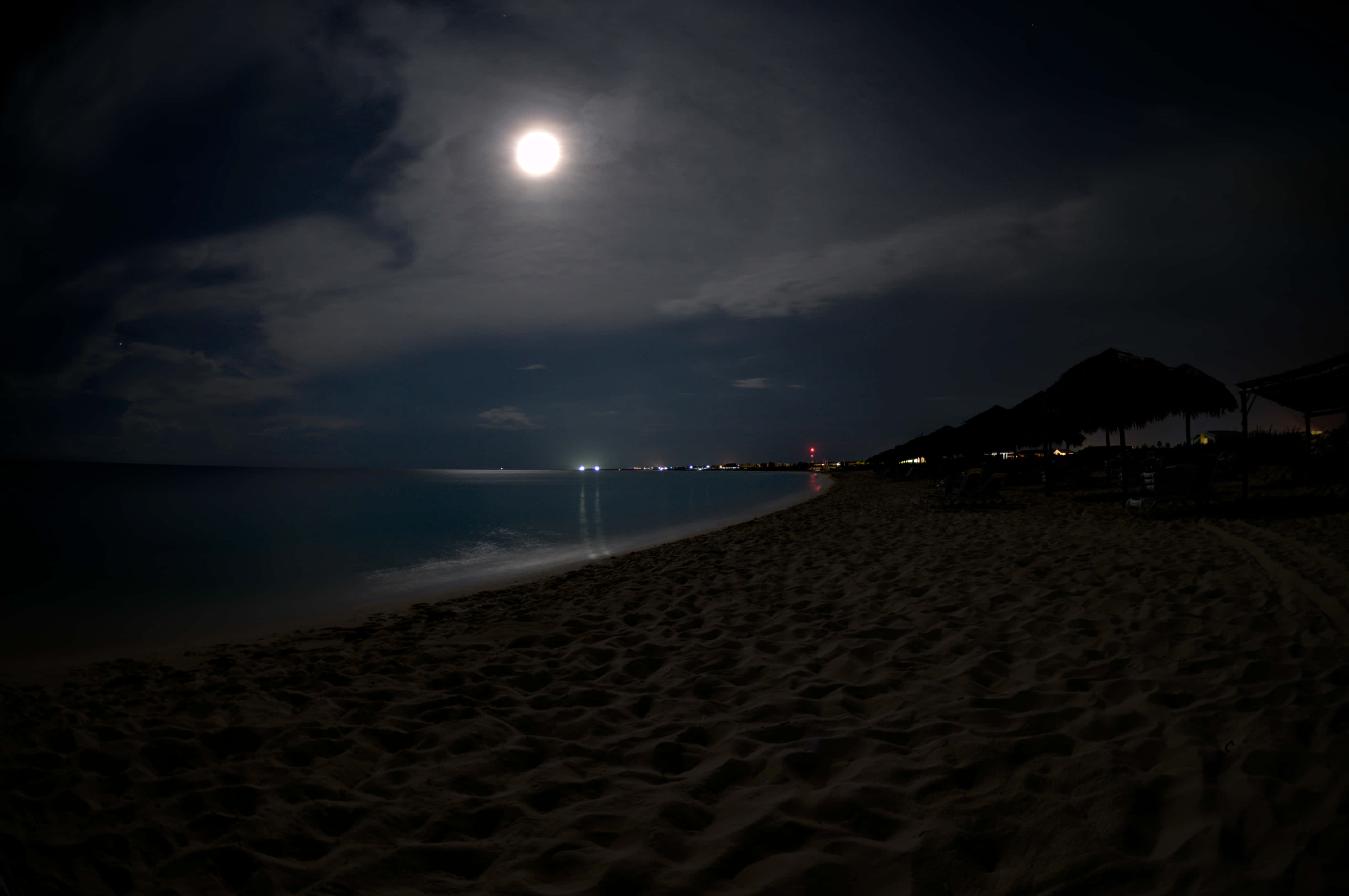 夜晚 桌布 壁紙 — 一輪明亮的滿月照亮了夜晚寧靜的海灘，伴隨著烏雲和沿岸遠處的燈光。