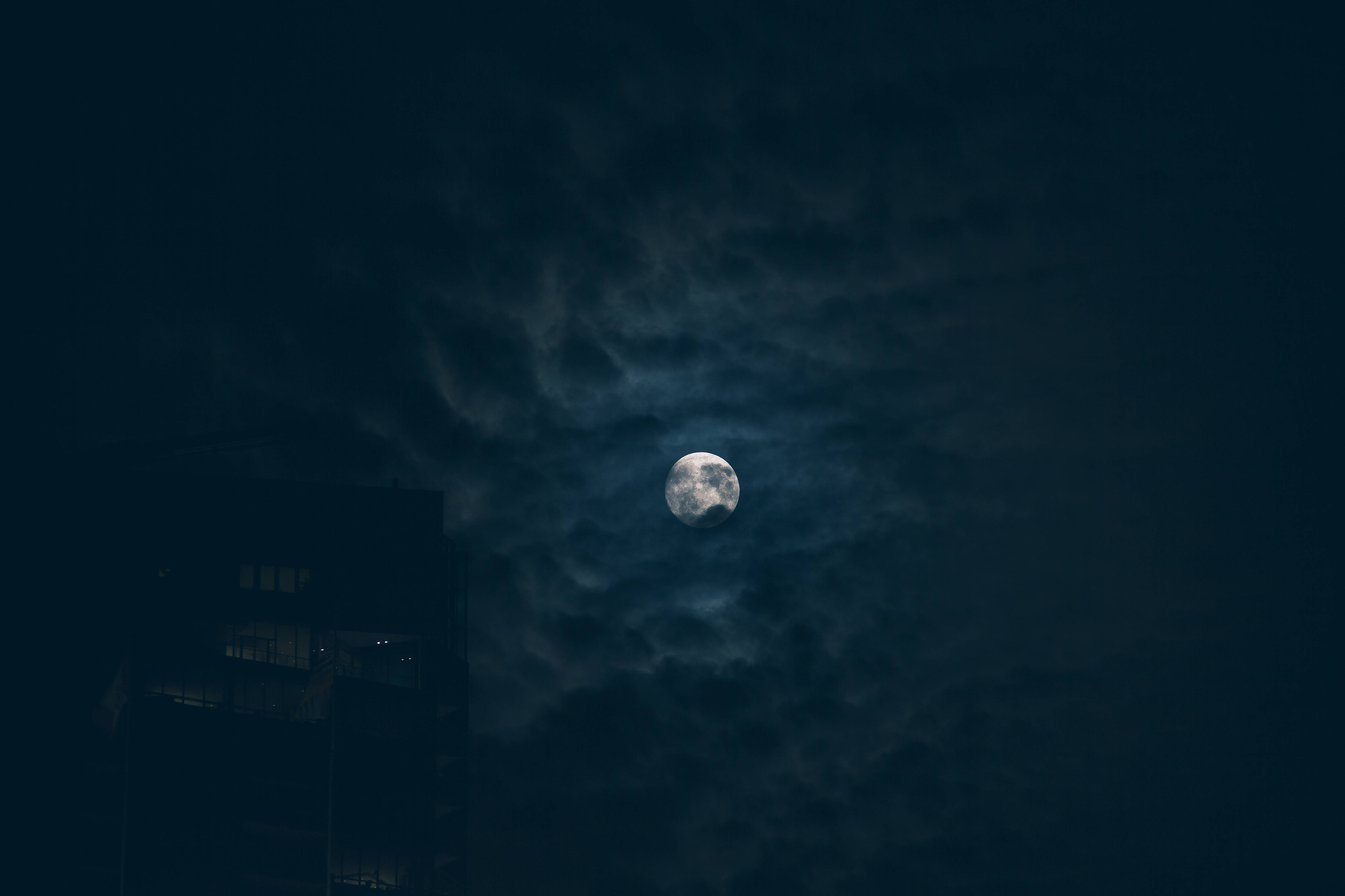 月光 桌布 壁紙 — 一輪明亮的滿月透過輕薄的雲層，在夜晚黑暗的城市天際線上發光。