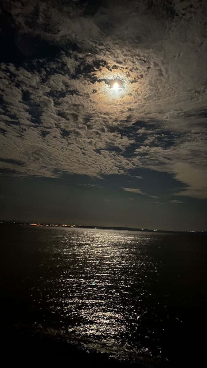 夜空 桌布 壁紙 — 一輪明月透過薄雲照耀，在黑暗的水面上反射其光芒，地平線上有遠處的城市燈光。