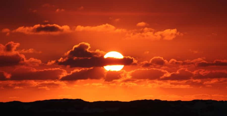 Clouds Aesthetics 桌布 壁紙 — 一輪火紅的夕陽在戲劇性的雲層後落下，在黑暗的剪影地平線上投下溫暖的光芒。