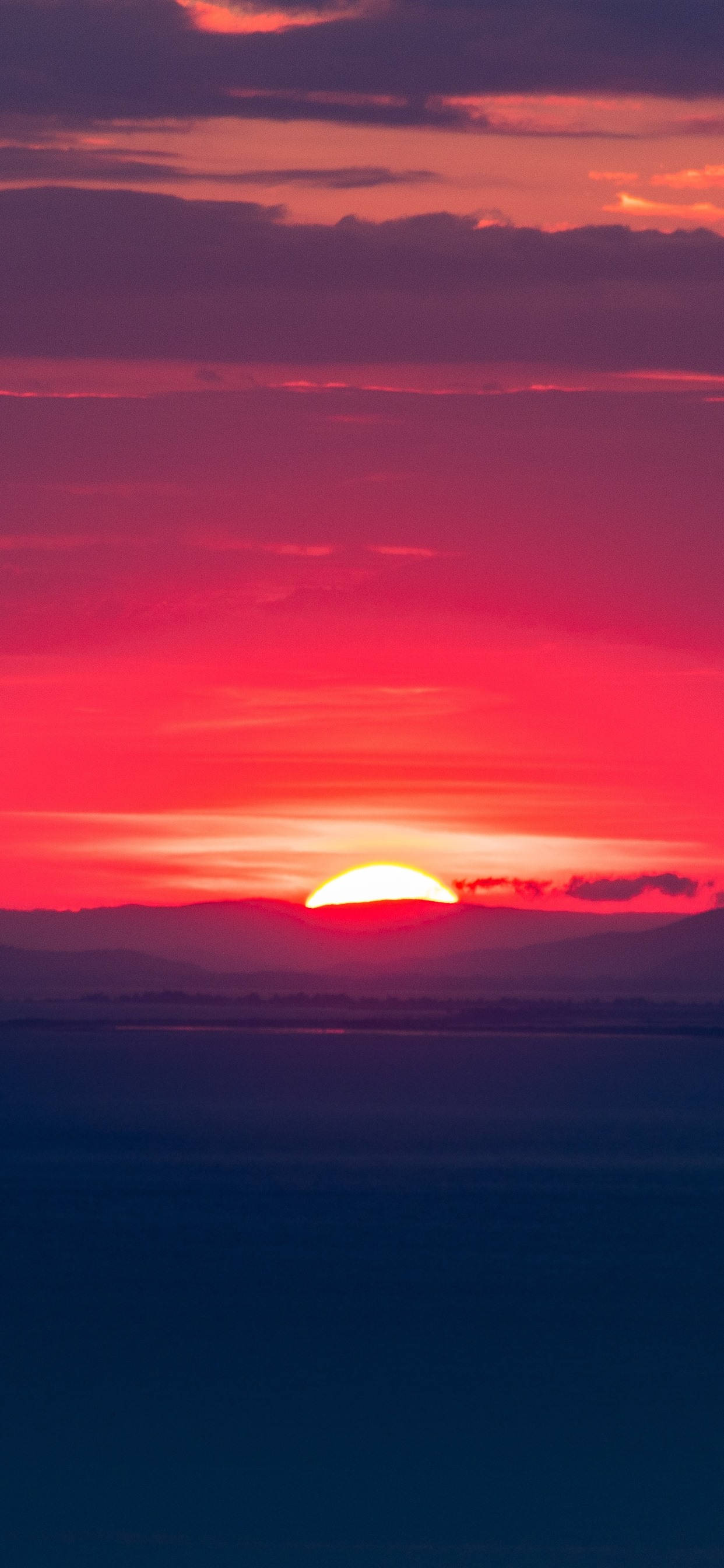 夕陽天空 桌布 壁紙 — 一輪火紅的夕陽落在遠山之後，在平靜的海平線上投下鮮豔的色彩。