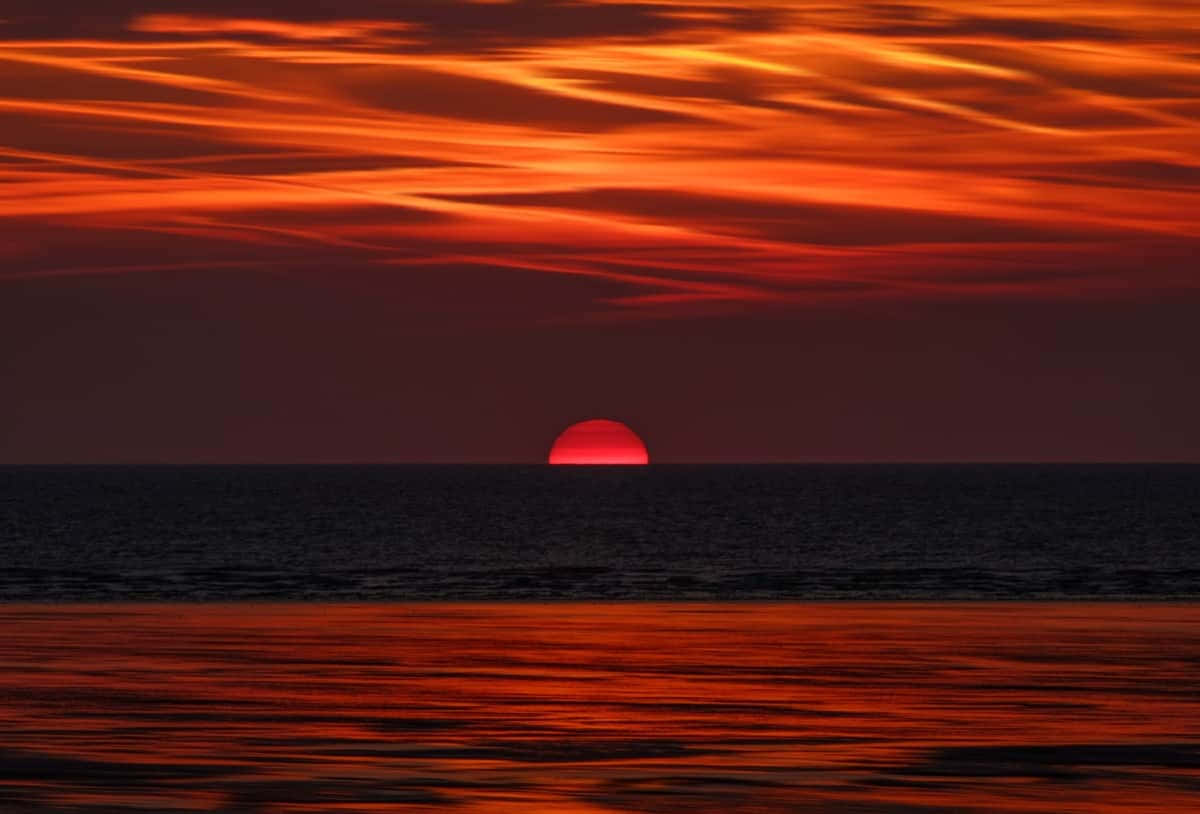 Orange Sunset 桌布 壁紙 — 一輪火紅的太陽在平靜的海平面上緩緩落下，映照出鮮豔的橙色和深紅色的雲彩。