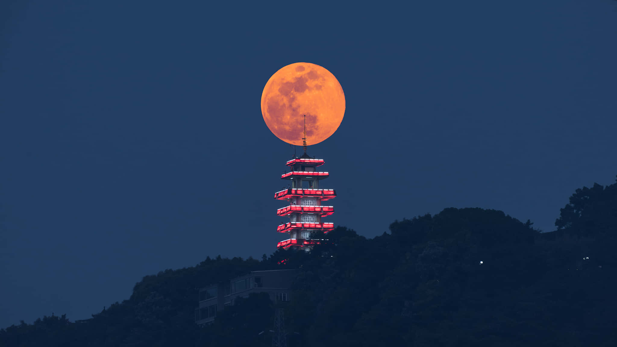 Pink Moon 桌布 壁紙 — 一輪發光的橙色超級月亮完美地與黑暗山頂上燈火通明的寶塔對齊，在黃昏時分。