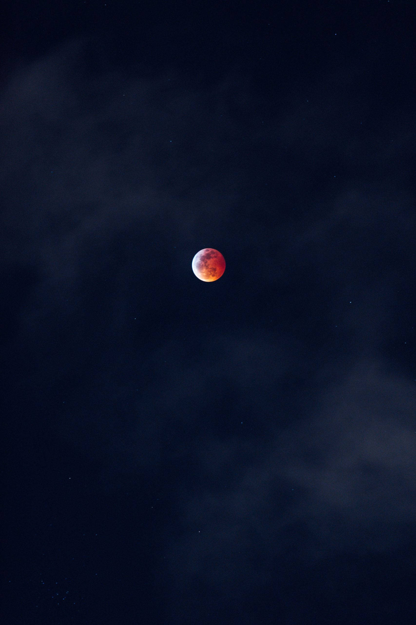 Red And Black iPhone 桌布 壁紙 — 一輪血紅色的月亮在佈滿遙遠星點的黑暗多雲夜空中微弱地發光。