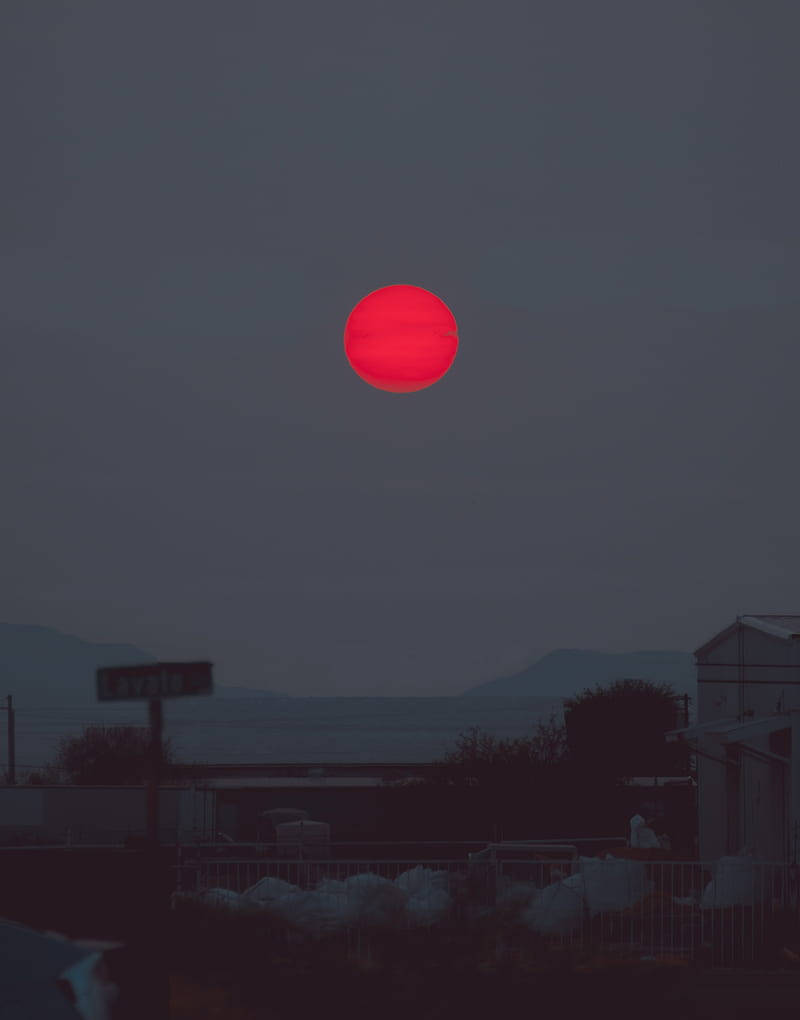 詭異 桌布 壁紙 — 一輪鮮紅的太陽低垂在黃昏的海邊小鎮上空，在遠山和屋頂上投下陰鬱的光芒。