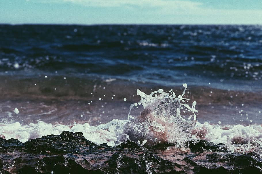 海灘 桌布 壁紙 — 一道充滿動感的海洋波浪衝擊著深色岩石，在蒼白的天空下激起白色的泡沫飛濺到空中。
