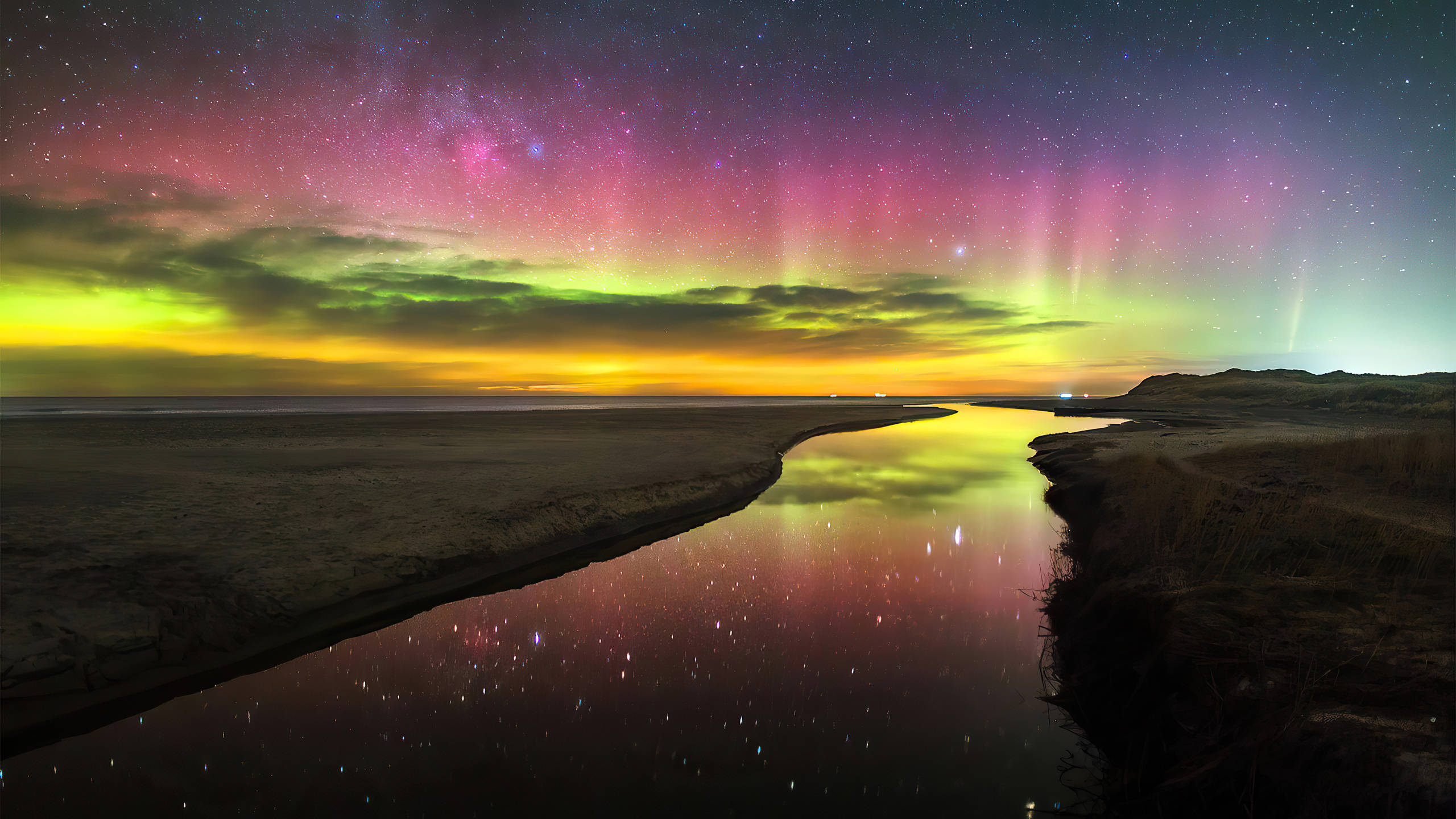 2560x1440 nature 桌布 壁紙 — 一道充滿活力的北極光在繁星點點的夜空下舞動，映照在一條平靜的河流上，反射出天體的光芒。