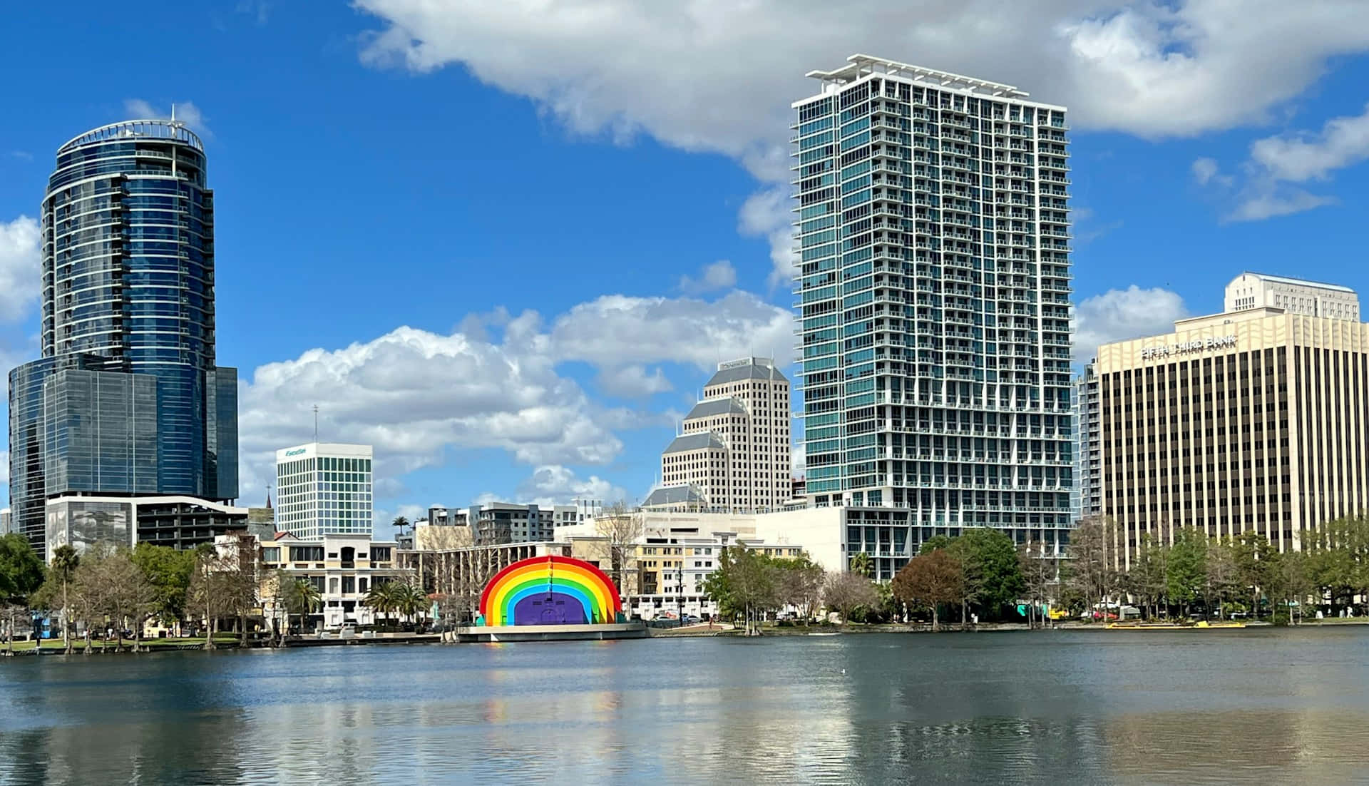 Orlando 桌布 壁紙 — 一道充滿活力的彩虹拱門漂浮在平靜的湖面上，倒映著現代摩天大樓，背景是明亮的藍天和蓬鬆的白雲。