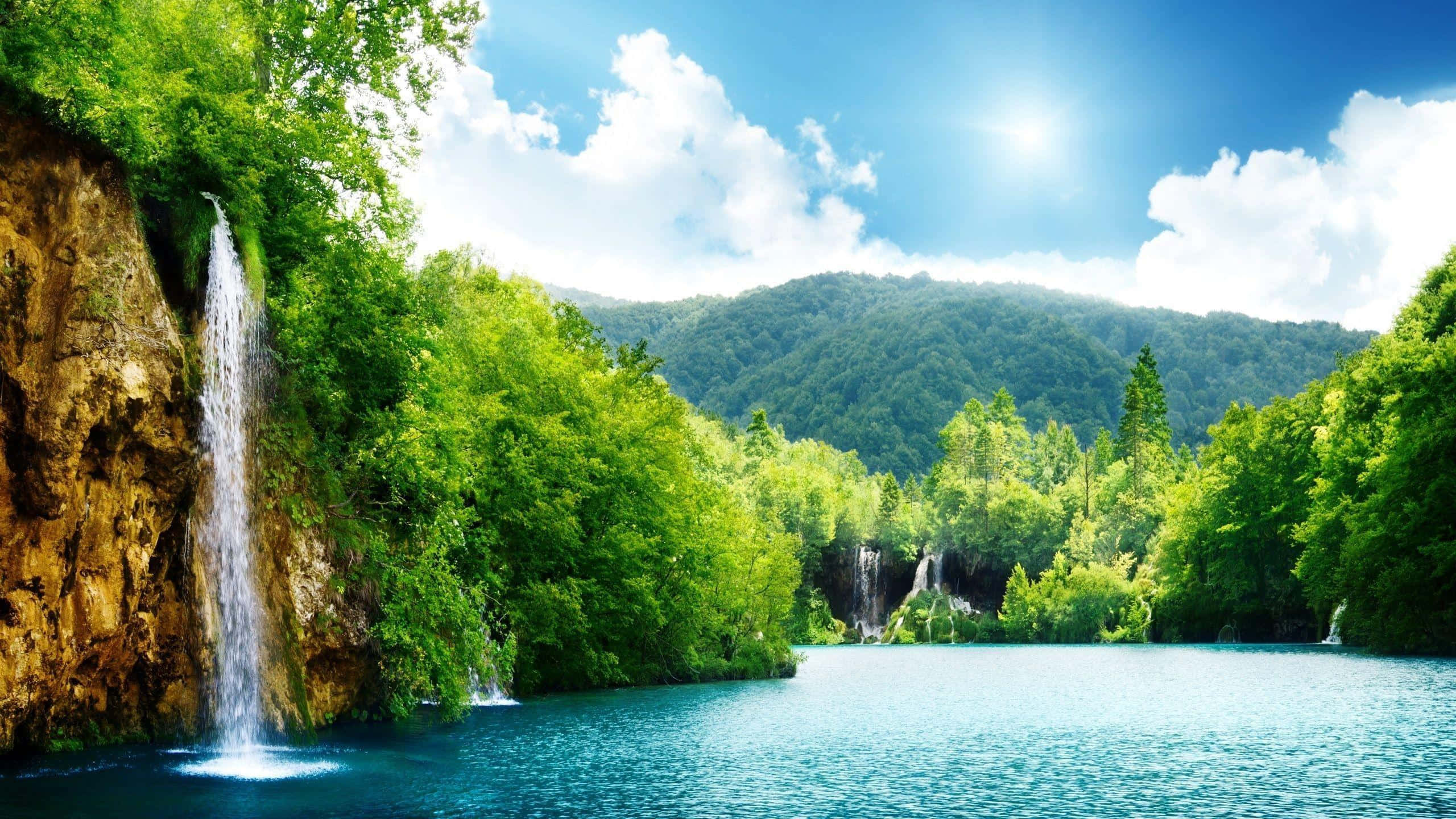 Relaxing Desktop 桌布 壁紙 — 一道充滿活力的瀑布傾瀉而下，流入一個被茂密綠色森林和遠處連綿起伏的丘陵環繞的碧綠湖泊，上方是明亮的藍天。