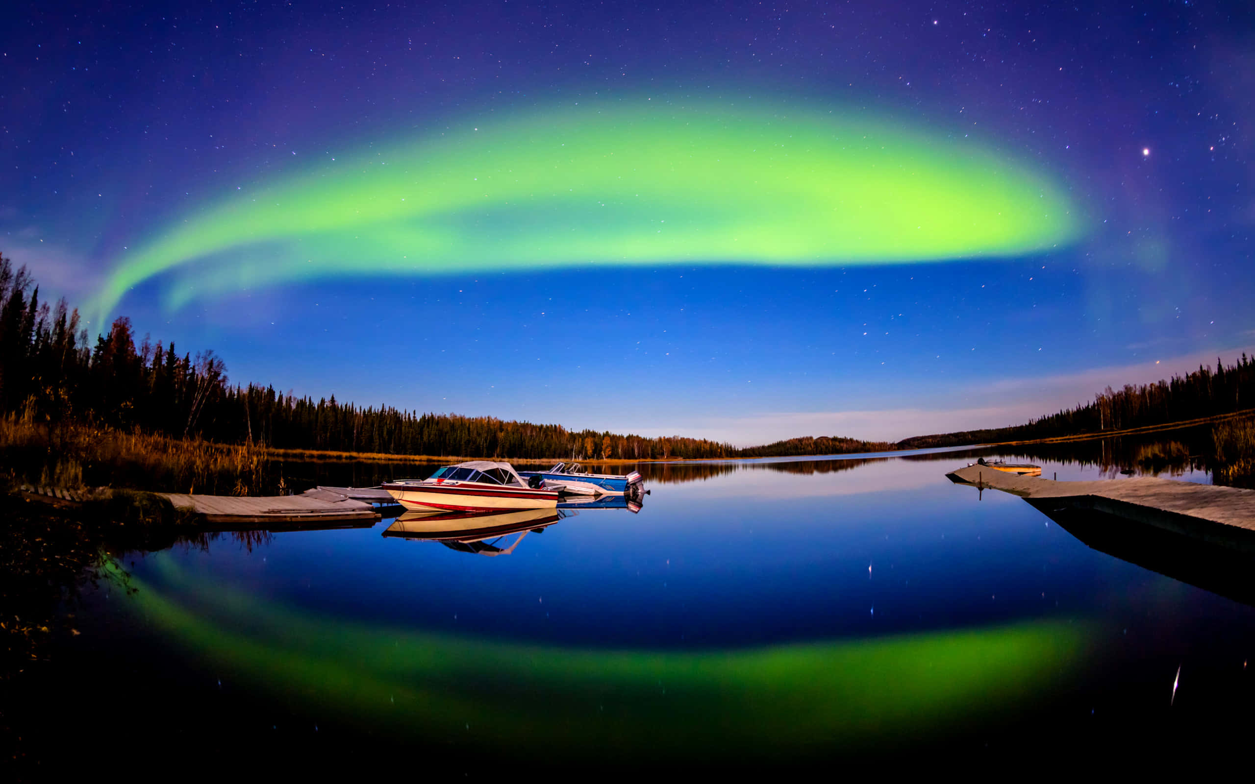aurora borealis 桌布 壁紙 — 一道充滿活力的綠色極光拱形橫跨繁星點點的夜空，完美倒映在平靜的湖面上，旁邊有停泊的船隻和樹木繁茂的湖岸。