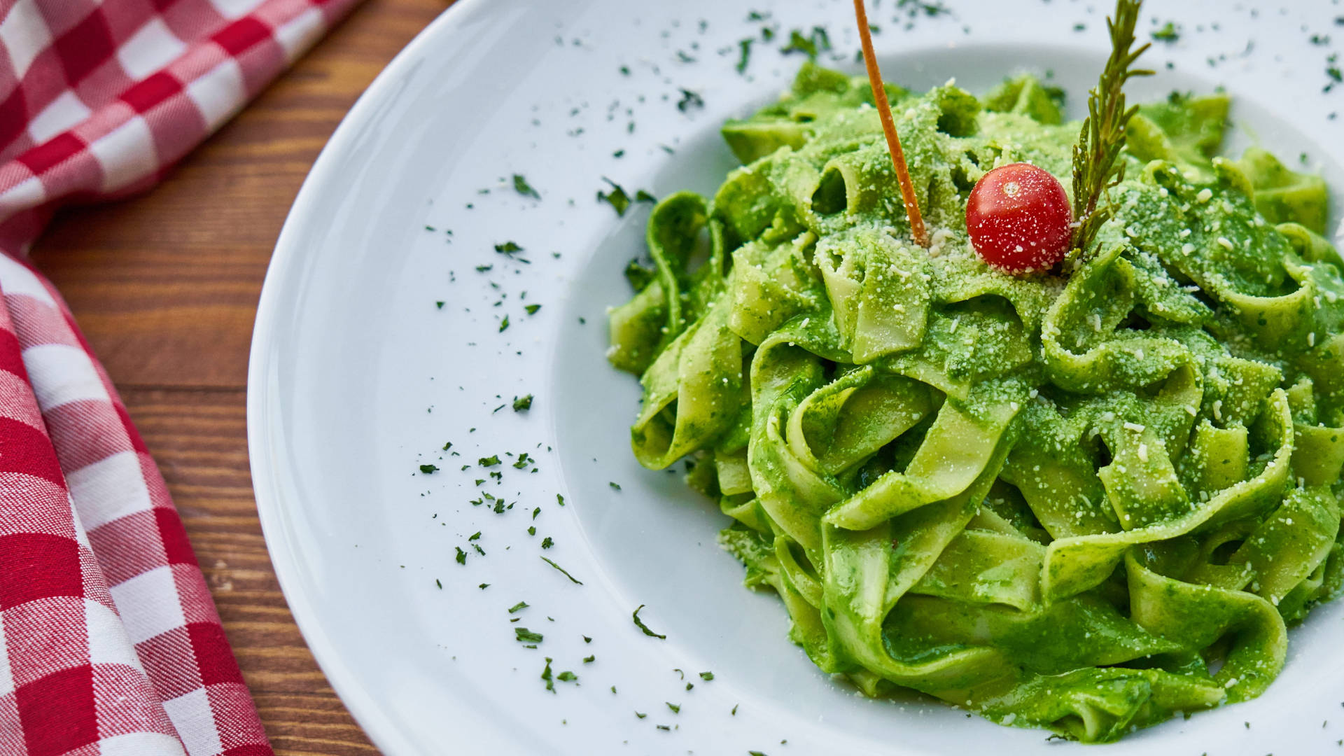 Pasta 桌布 壁紙 — 一道充滿活力的綠色青醬義大利麵，以櫻桃番茄和新鮮香草點綴，放在白色盤子上，旁邊有一張紅色格子餐巾。