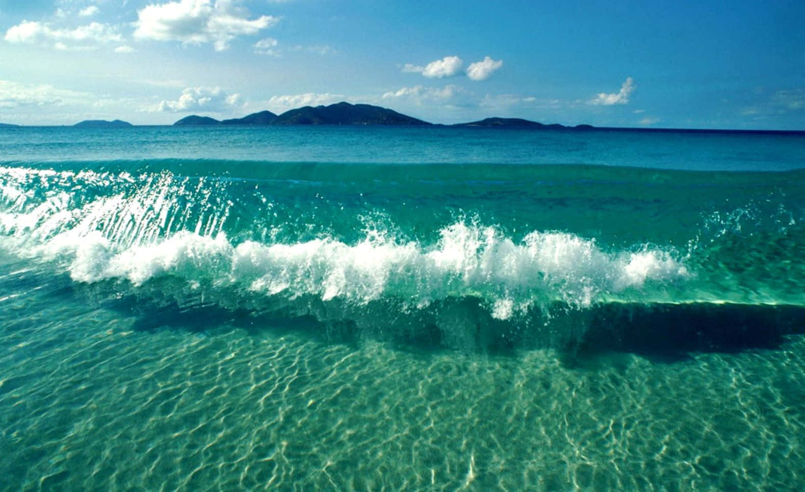 waves 桌布 壁紙 — 一道充滿活力的藍綠色海浪帶著白色泡沫衝過清澈的淺水，朝著明亮的藍天下遠處朦朧的島嶼翻騰。
