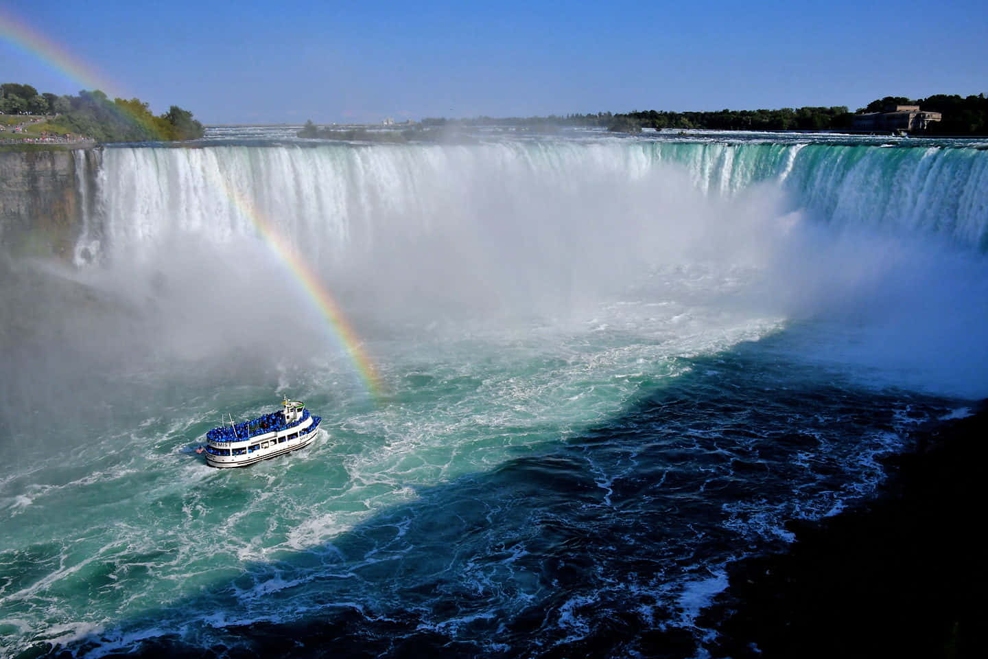 Niagara Falls 桌布 壁紙 — 一道壯麗的彩虹橫跨在轟鳴的尼亞加拉大瀑布之上，一艘觀光船在下方湍急的碧綠海水中航行。