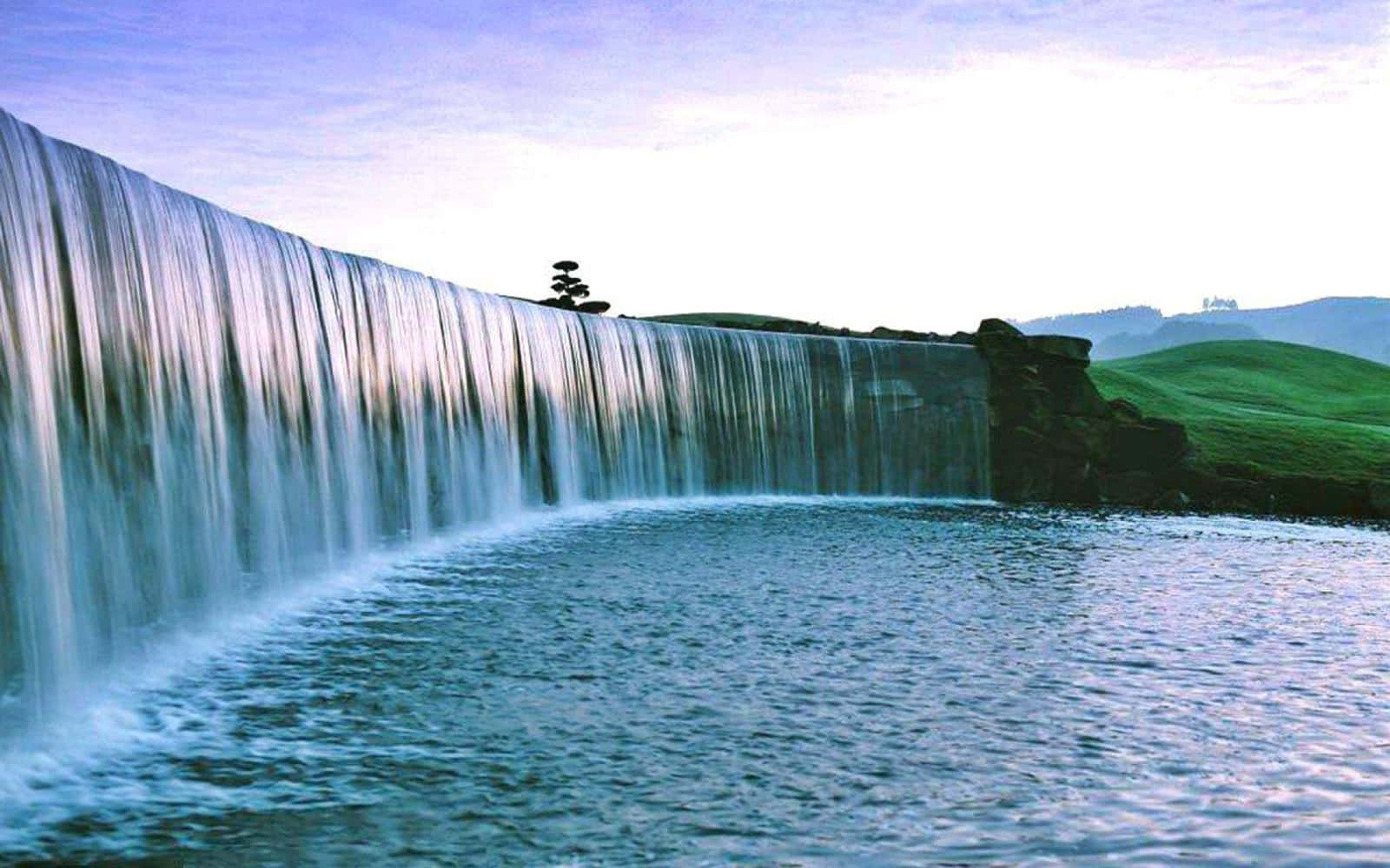 風景動畫 桌布 壁紙 — 一道寧靜的寬闊瀑布流入平靜的水池，背景是鬱鬱蔥蔥的綠色山丘和柔和的紫色天空。