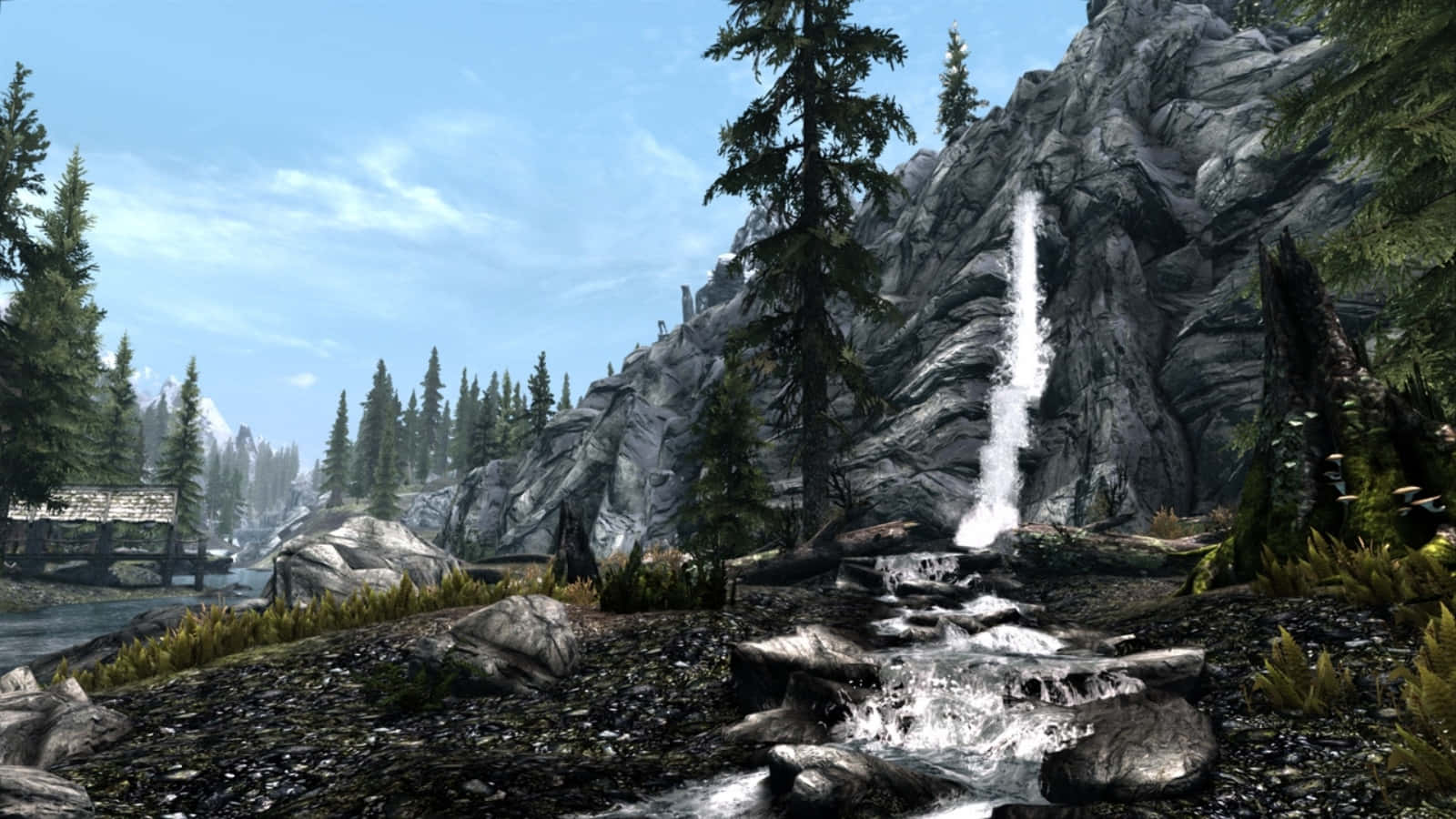 Skyrim Landscapes 桌布 壁紙 — 一道寧靜的山間瀑布從灰色懸崖傾瀉而下，流入岩石溪流，四周環繞著常綠樹木，在清澈的藍天下。