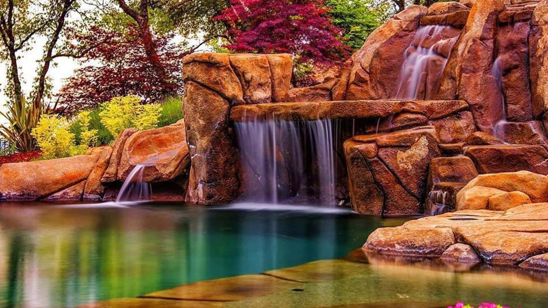 Beautiful Place 桌布 壁紙 — 一道寧靜的瀑布從崎嶇的棕色岩石上傾瀉而下，流入一個被色彩繽紛的秋葉環繞的平靜碧綠水池。