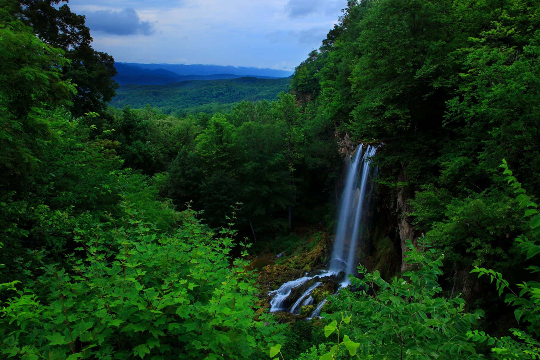 Blue Ridge Mountains 桌布 壁紙 — 一道寧靜的瀑布穿過茂密的綠色森林，遠山在陰天之下。