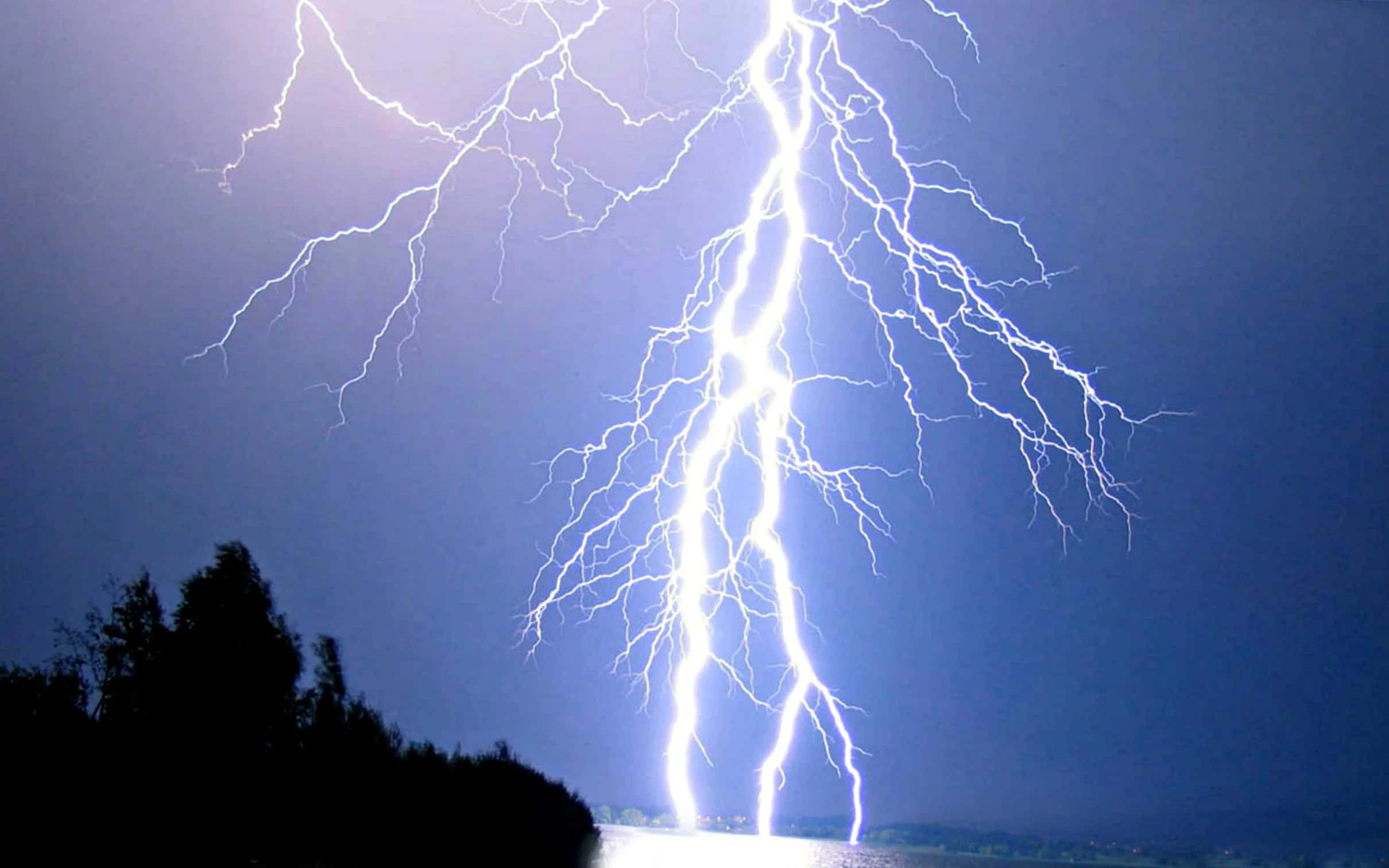 Lightning Bolt 桌布 壁紙 — 一道巨大的叉狀閃電照亮了暗藍色的夜空，下方是樹木的剪影和反射的水面。