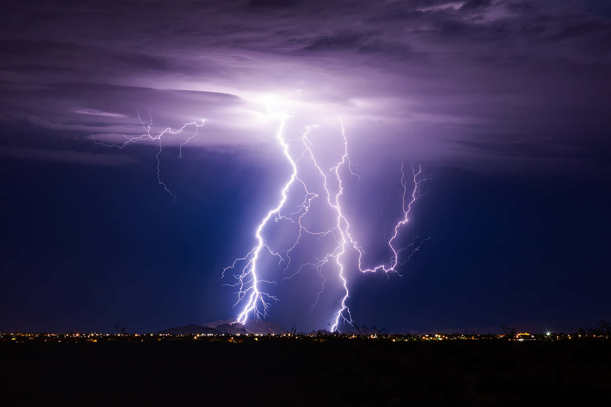 閃電 桌布 壁紙 — 一道巨大的紫色閃電劃破黑暗的暴風雨天空，下方是遠處的夜景城市天際線。