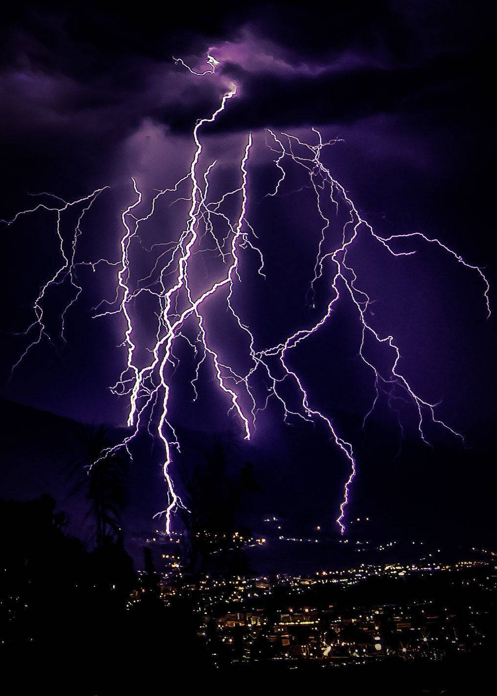 淡紫色 桌布 壁紙 — 一道巨大的紫色閃電在暴風雨的夜空中分叉，下方是剪影般的城市景觀，遠處燈光閃爍。