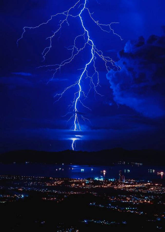 dark blue aesthetic tumblr 桌布 壁紙 — 一道巨大的藍色閃電在夜間劃過黑暗的海濱城市上空，照亮了水面和遠處的山丘。