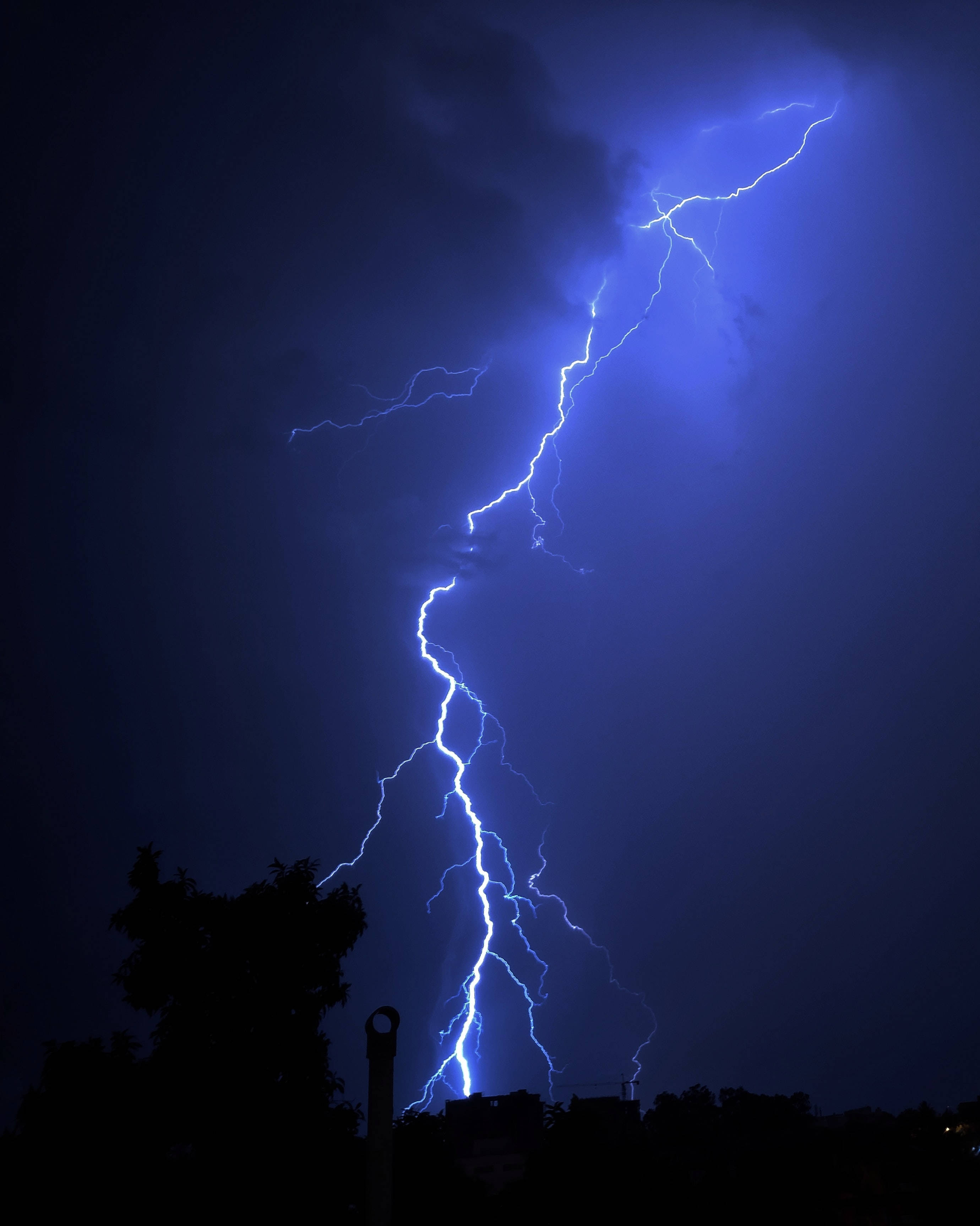 閃電 桌布 壁紙 — 一道巨大的藍色閃電在黑暗的暴風雨天空中分叉，掠過剪影般的樹木和建築物。