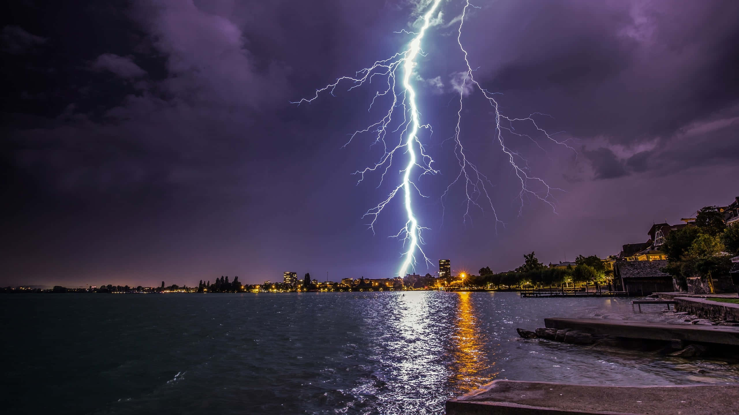暴風 桌布 壁紙 — 一道巨大的閃電在夜間劃過黑暗的湖面，照亮了湖水與遠處的城市燈光。