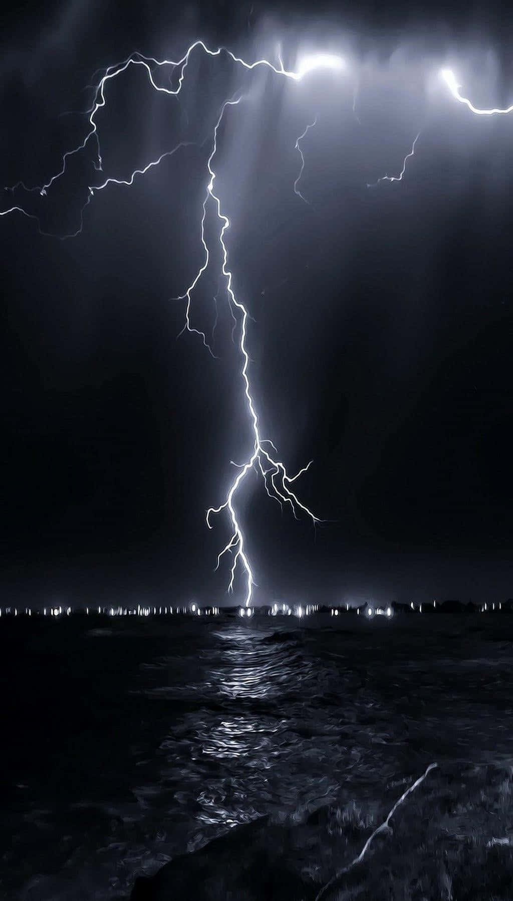 Black And White Storm 桌布 壁紙 — 一道巨大的閃電在夜間擊中海洋，照亮了黑暗的海浪和地平線上遙遠的城市燈光。