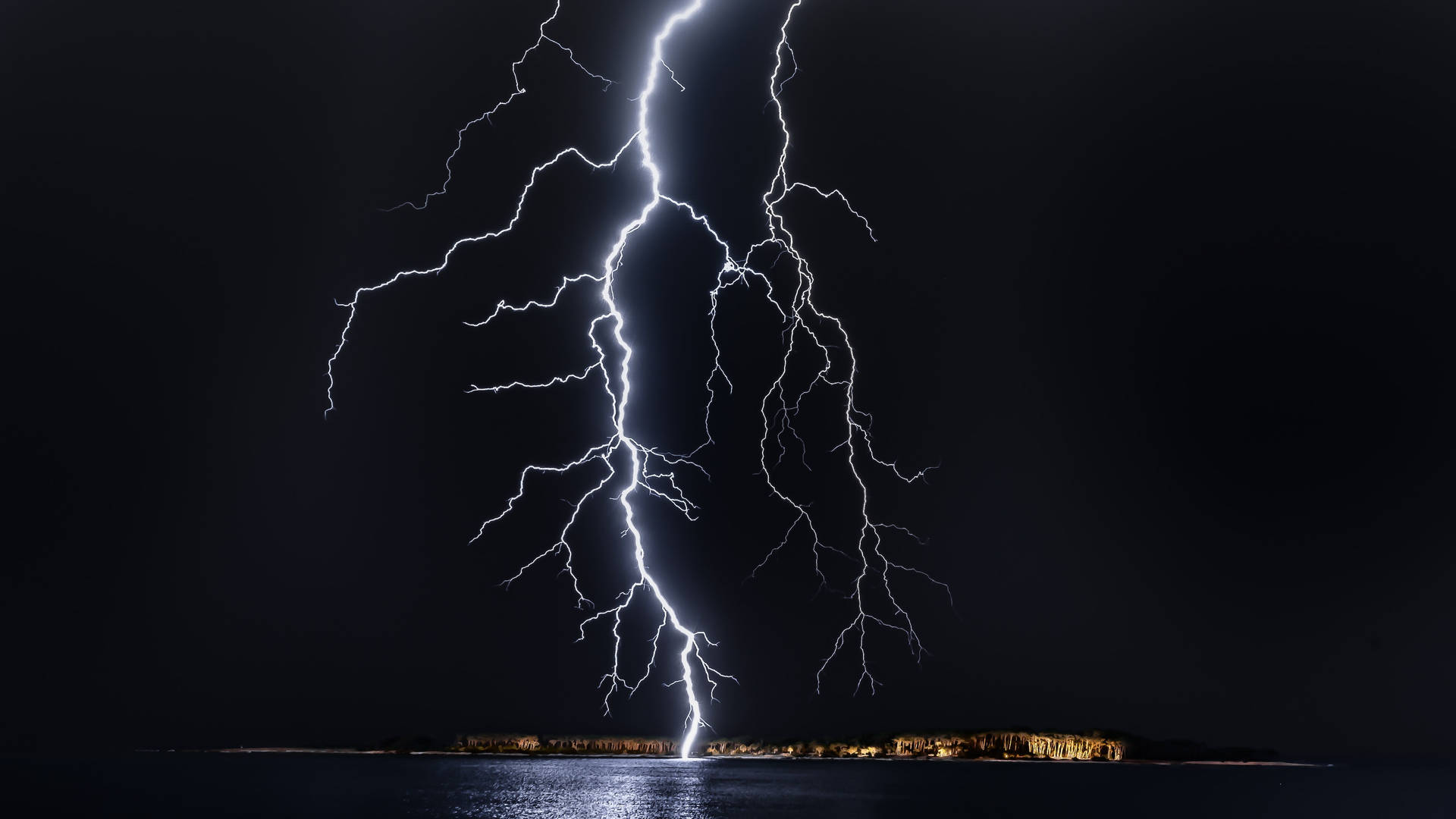  壁紙 — 一道巨大的閃電在平靜的水面上方的黑暗夜空中分叉，遠處有被照亮的海岸線。