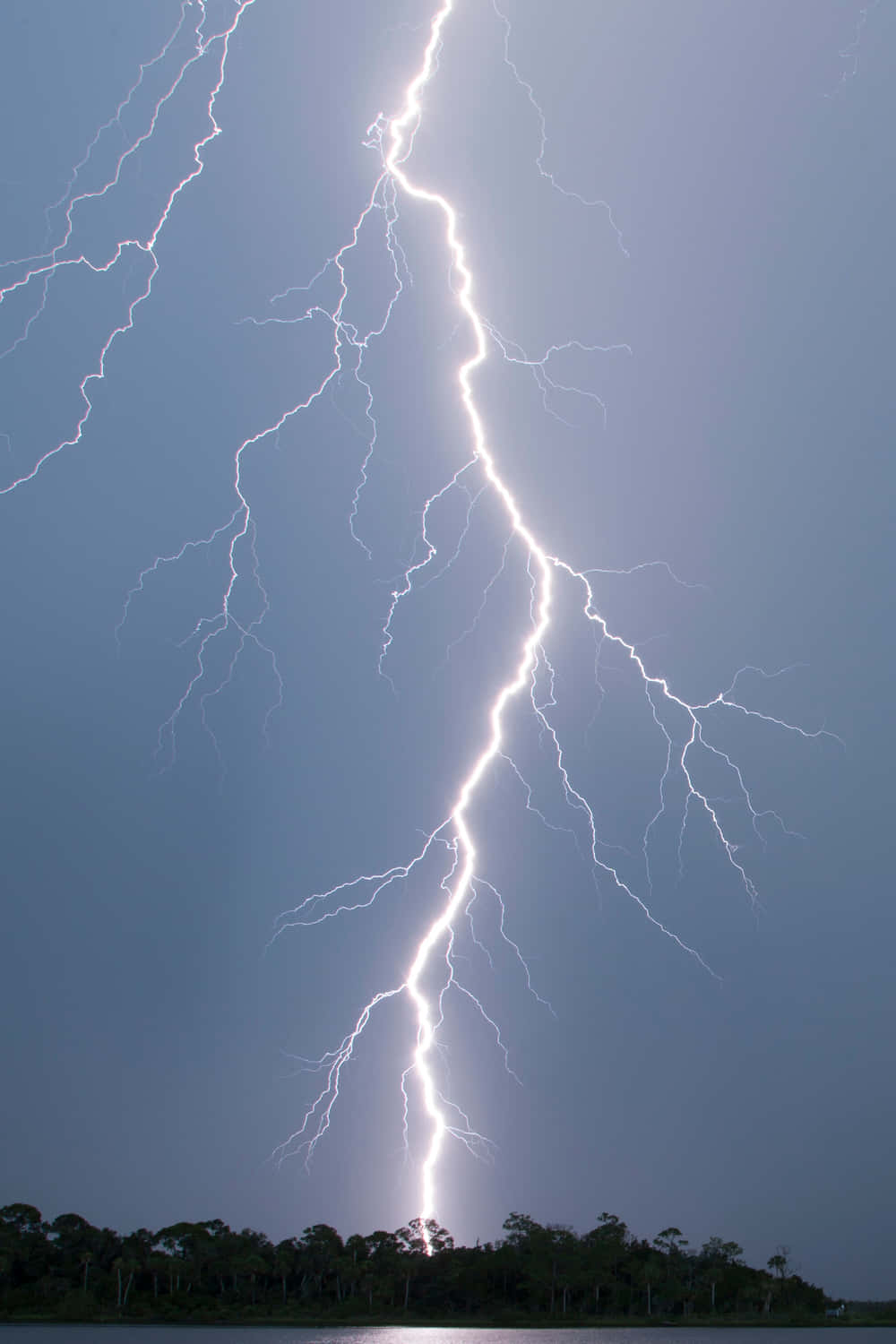 閃電 桌布 壁紙 — 一道巨大的閃電在平靜的湖泊和樹木成行的岸邊上方，劃過黑暗的暴風雨天空。