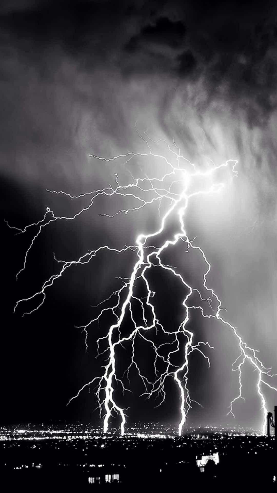 Black And White Storm 桌布 壁紙 — 一道巨大的閃電在暴風雨的夜空中分叉，照亮了遠處燈火輝煌的城市景觀。