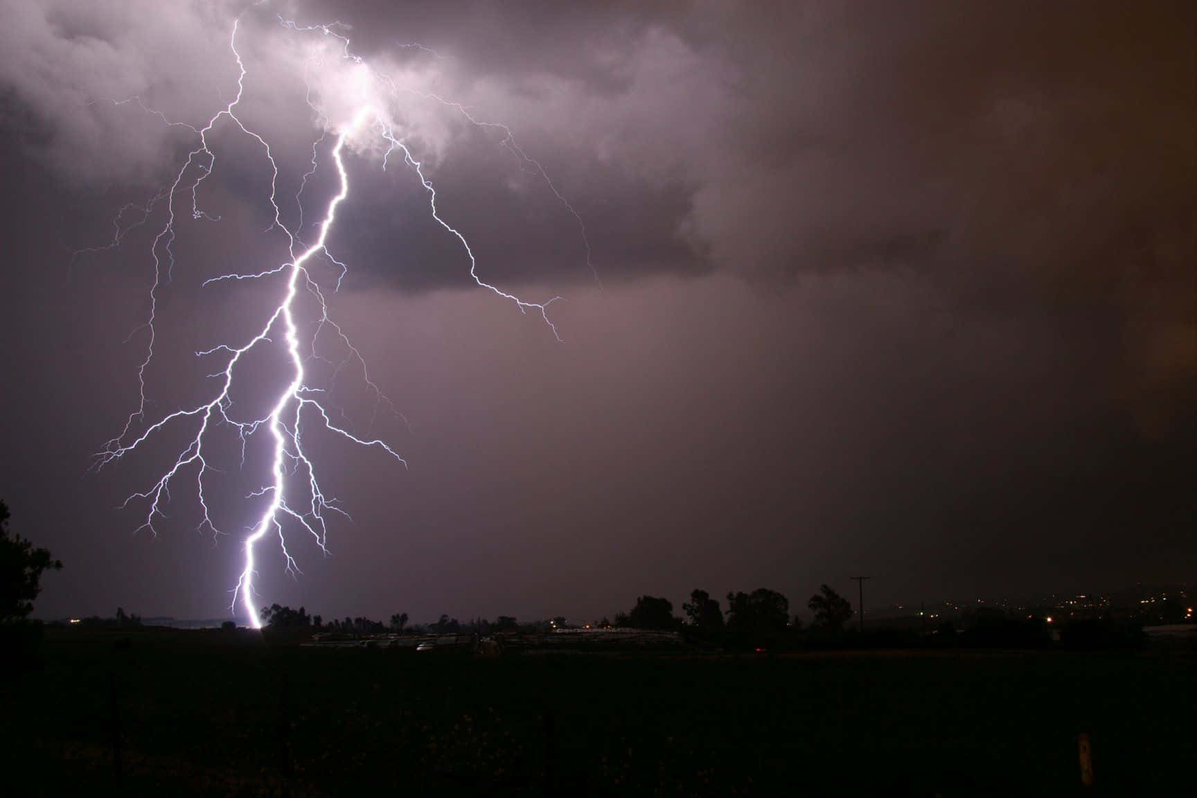 thunder 桌布 壁紙 — 一道巨大的閃電在暴風雨的夜空中分叉，下方是黑暗的鄉村景觀，遠處有城市燈光。