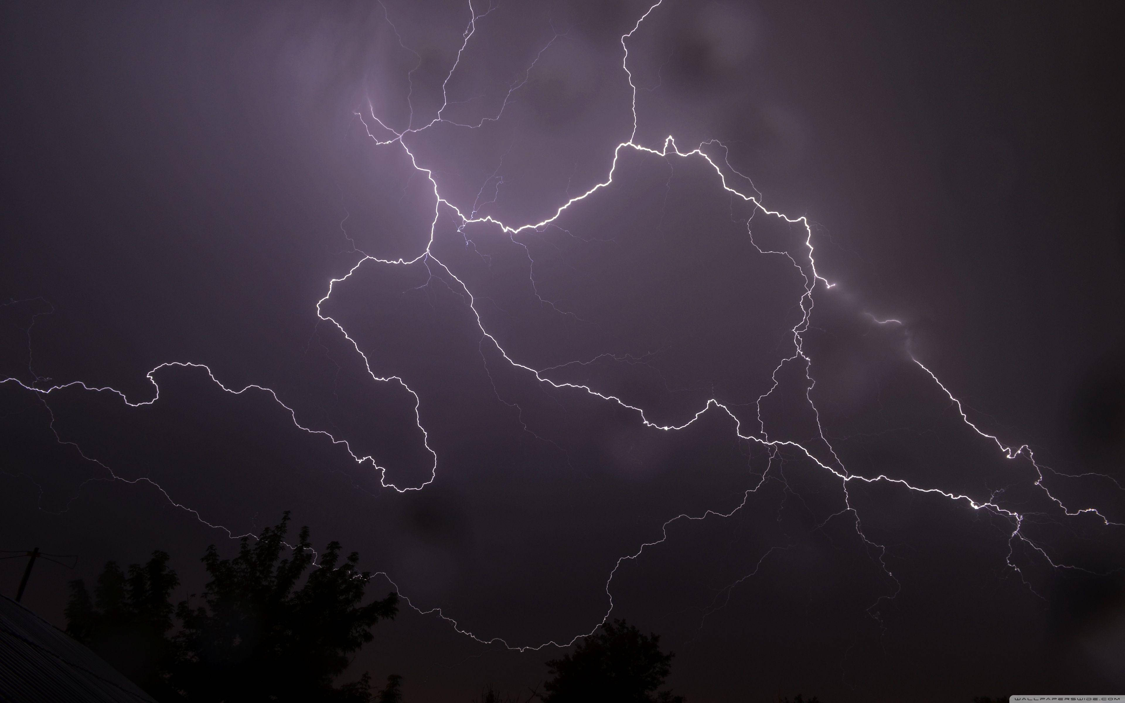 Aesthetic Lightning 桌布 壁紙 — 一道巨大的閃電在黑暗的暴風雨天空中分叉，籠罩在剪影般的樹木和屋頂之上。