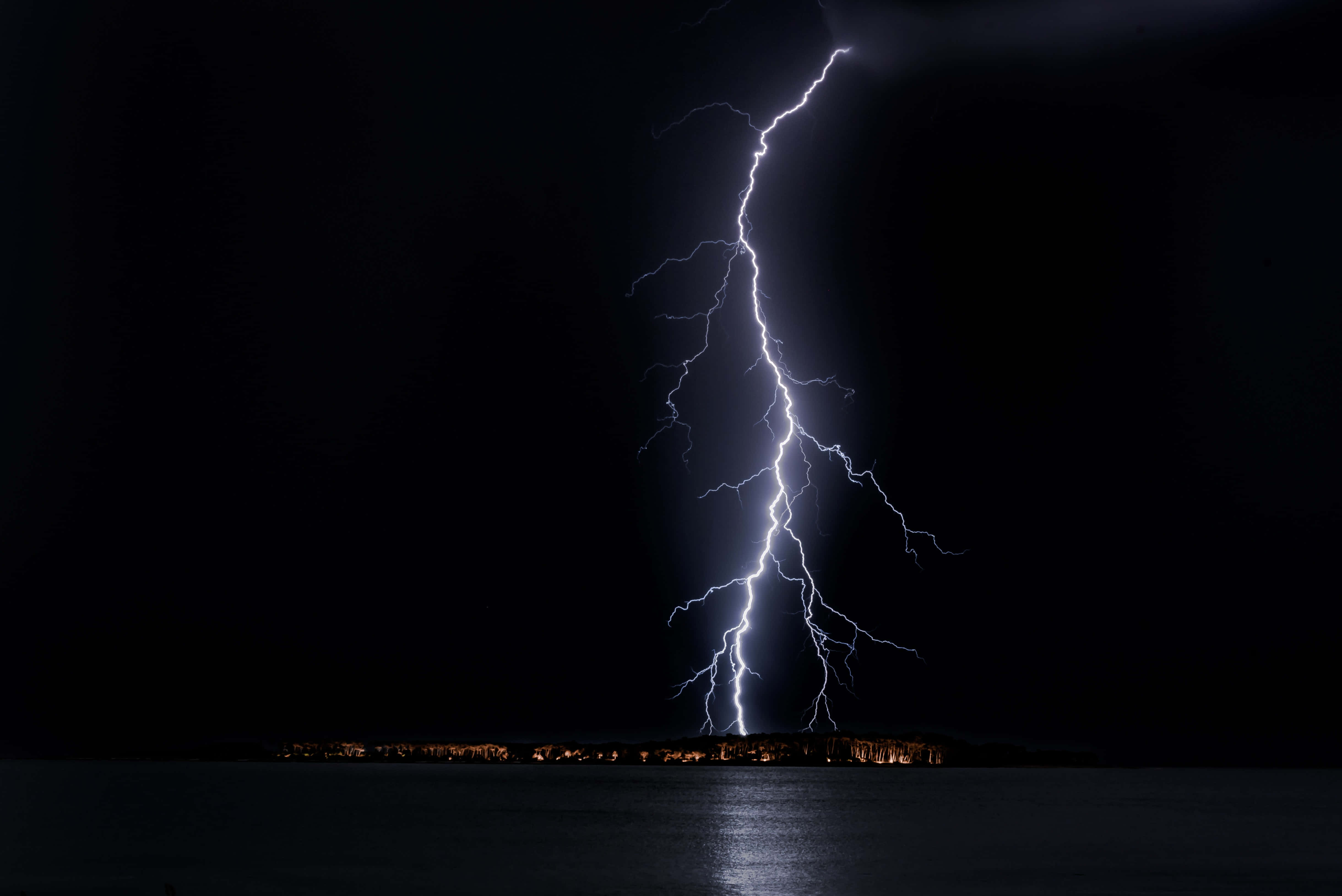 Lightning Bolt 桌布 壁紙 — 一道巨大的閃電擊落在黑暗的水面上，遠方的城市燈光在下方閃爍。