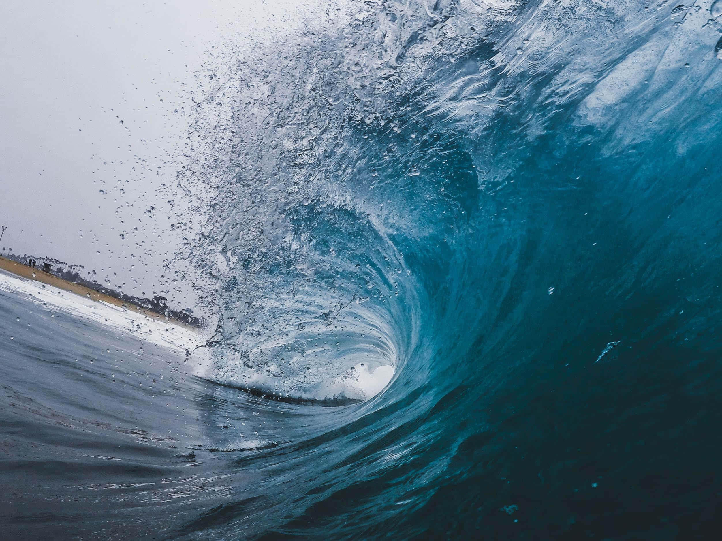 waves 桌布 壁紙 — 一道強勁的海浪捲成玻璃般的藍色隧道，帶有泡沫般的白色水花，映襯著灰色的天空。